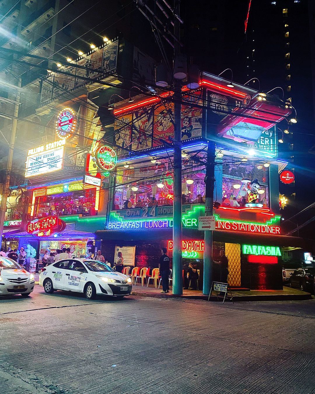 Brightly lit, multi-story building at night with colorful neon lights. Cars parked on street.