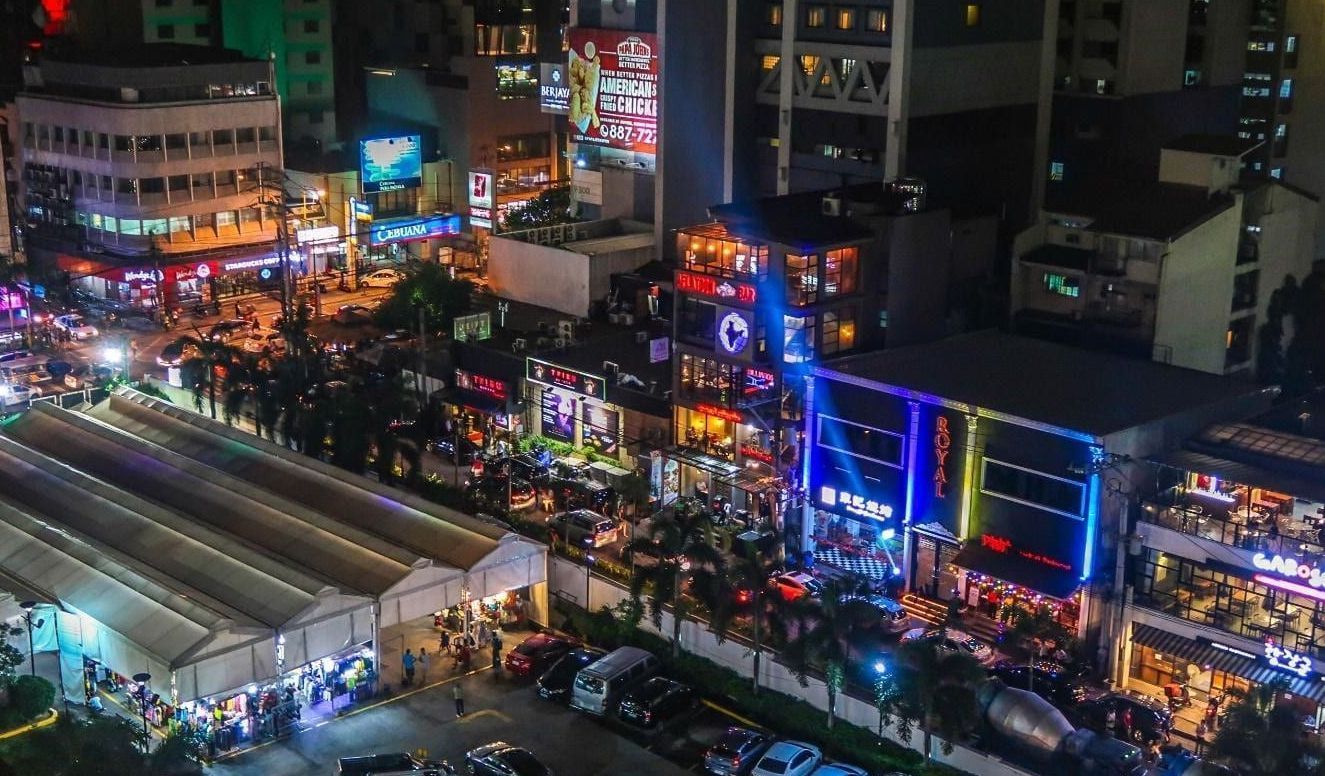 Night scene of a busy city street with lit-up buildings, people, cars, and tents.