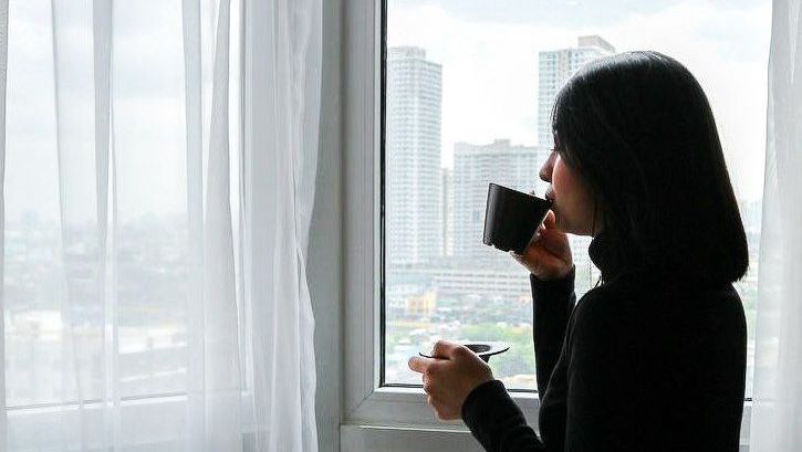 Woman sipping from a mug, looking out a window at city skyscrapers.
