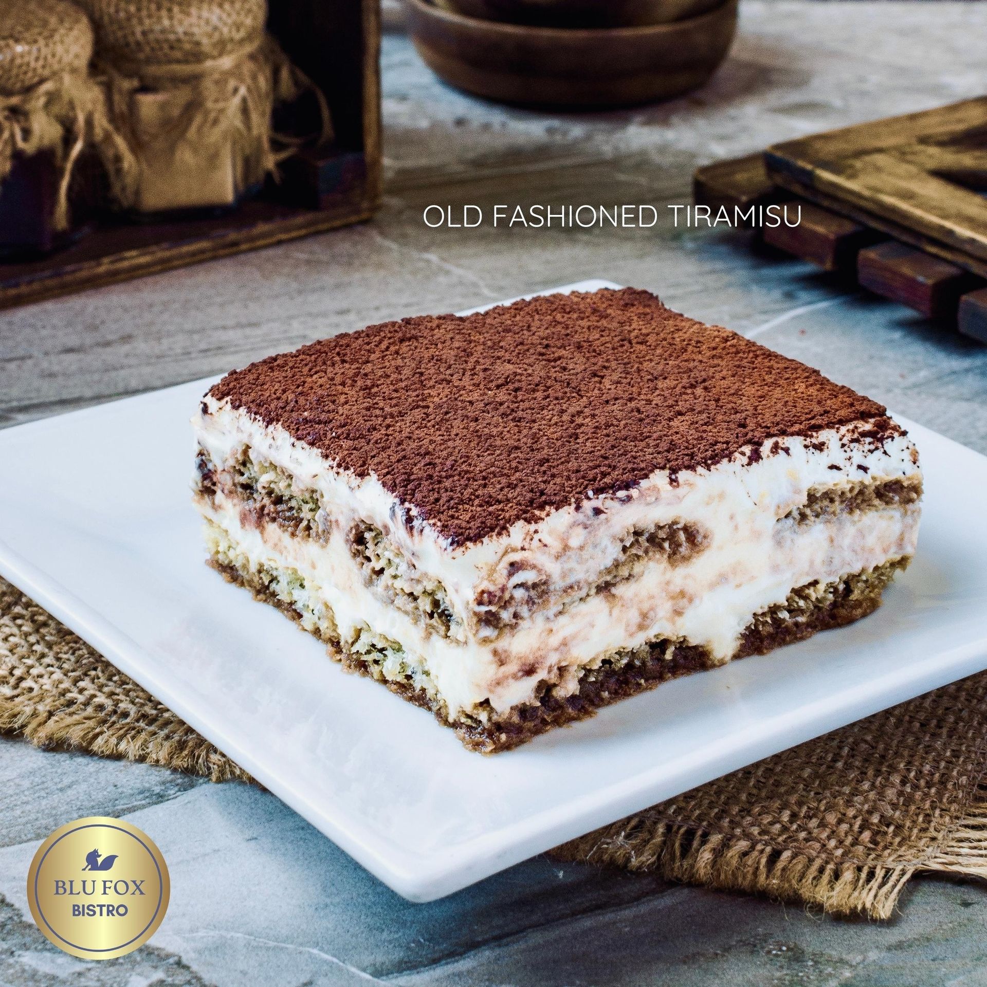 Old fashioned tiramisu on a white plate, dusted with cocoa powder, rustic setting.