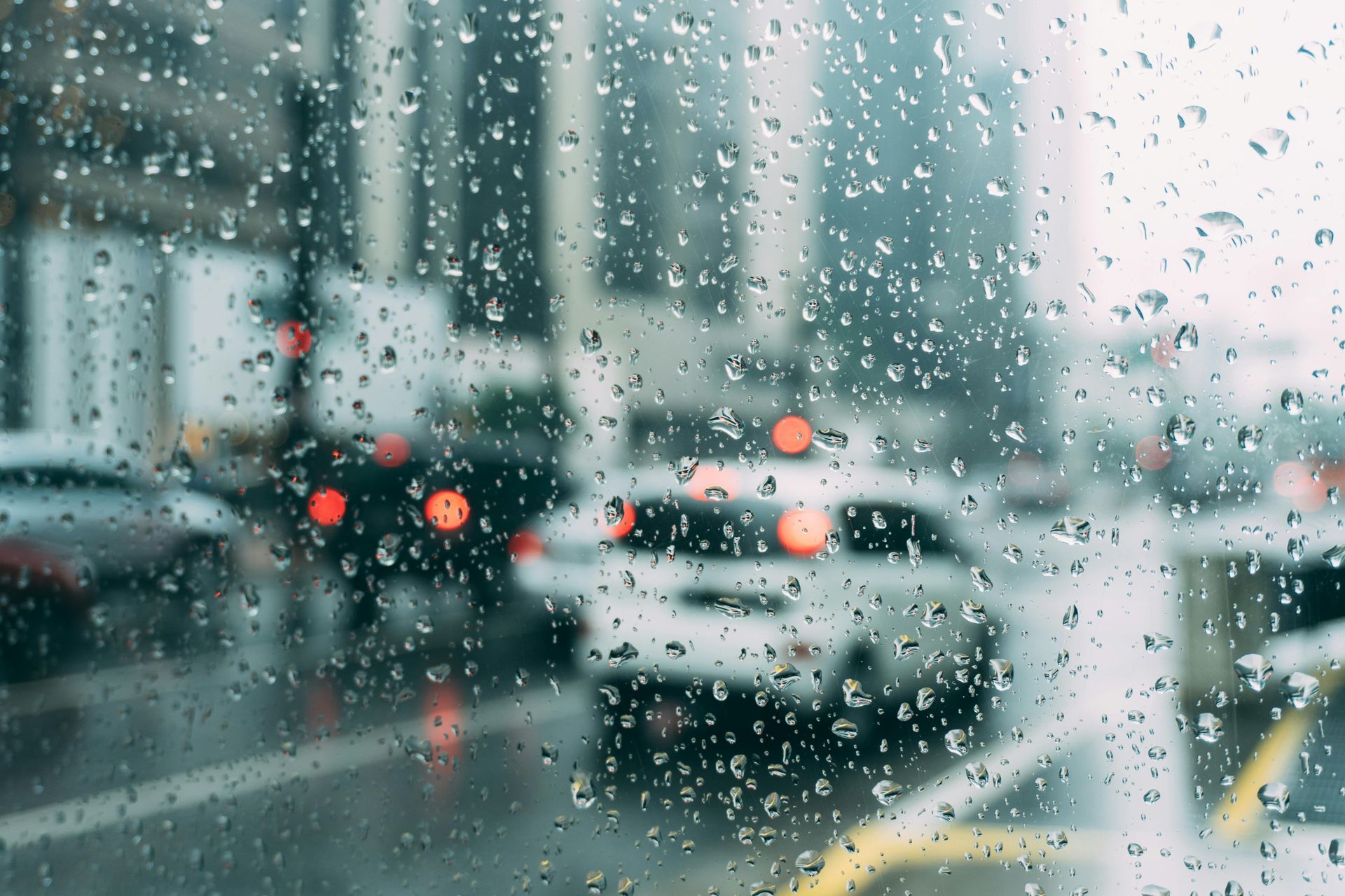Rainy city street seen through a wet window, with blurred cars and traffic lights.