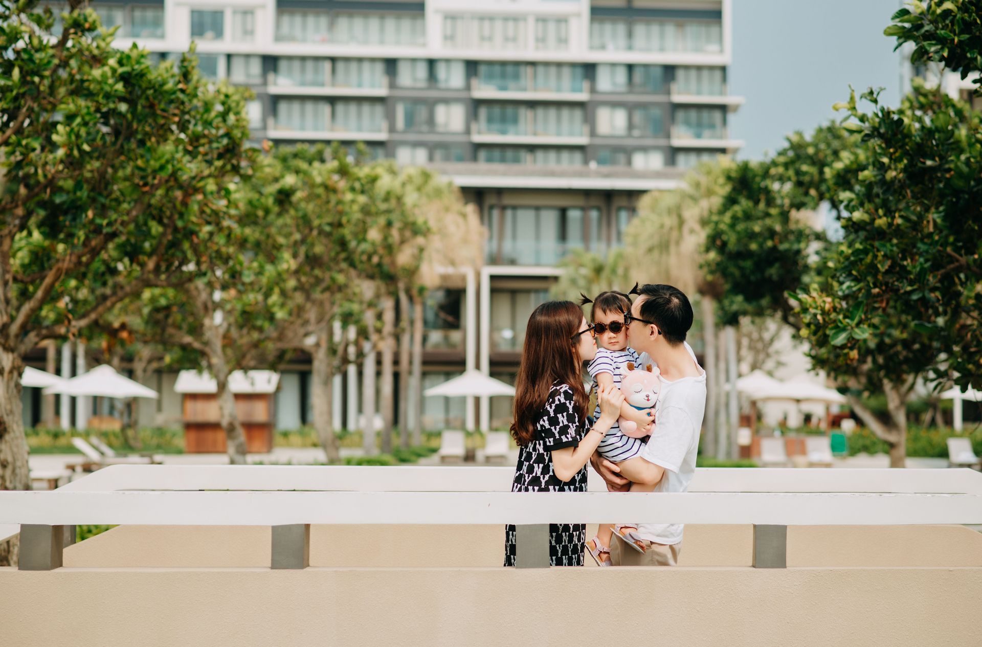 Couple holding a baby in front of a modern building and trees, outdoors.