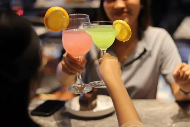 Two colorful cocktails with orange slices being toasted at a restaurant table