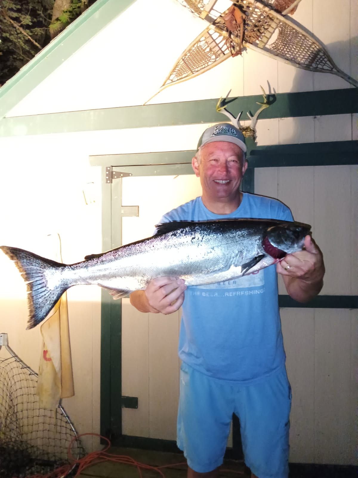 Man holding a large silver fish outdoors, wearing a blue shirt and shorts, smiling at the camera