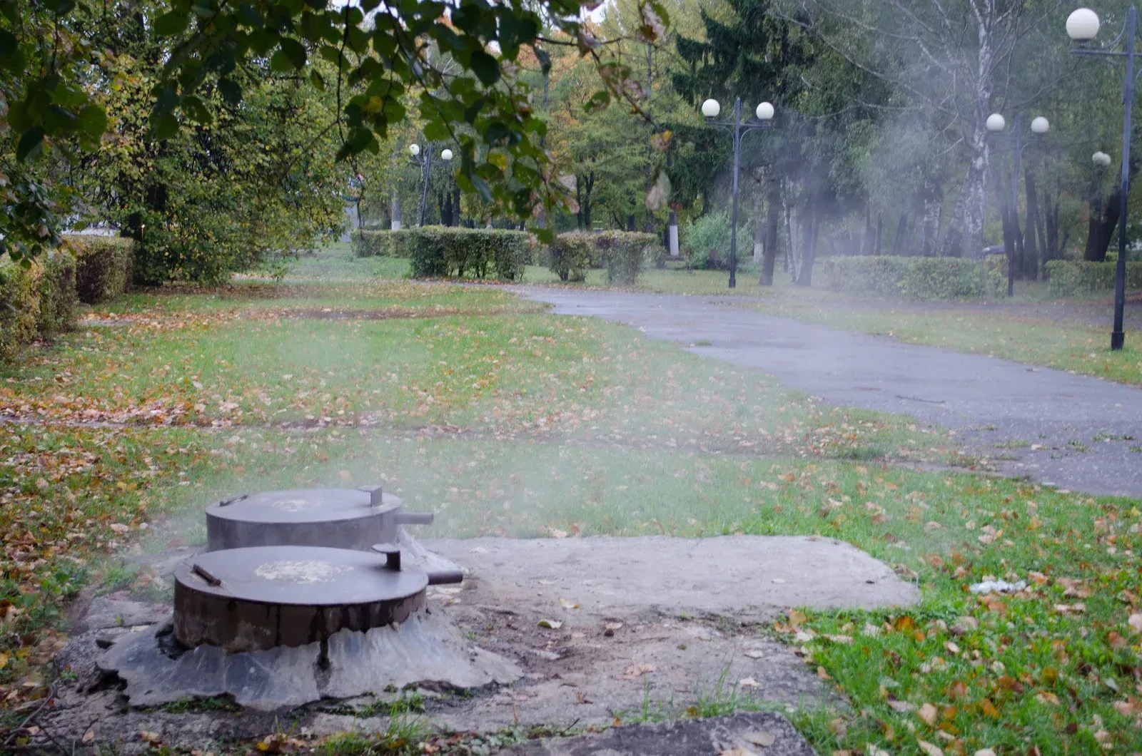 Two metal lids on concrete, misting in a park with trees and path.