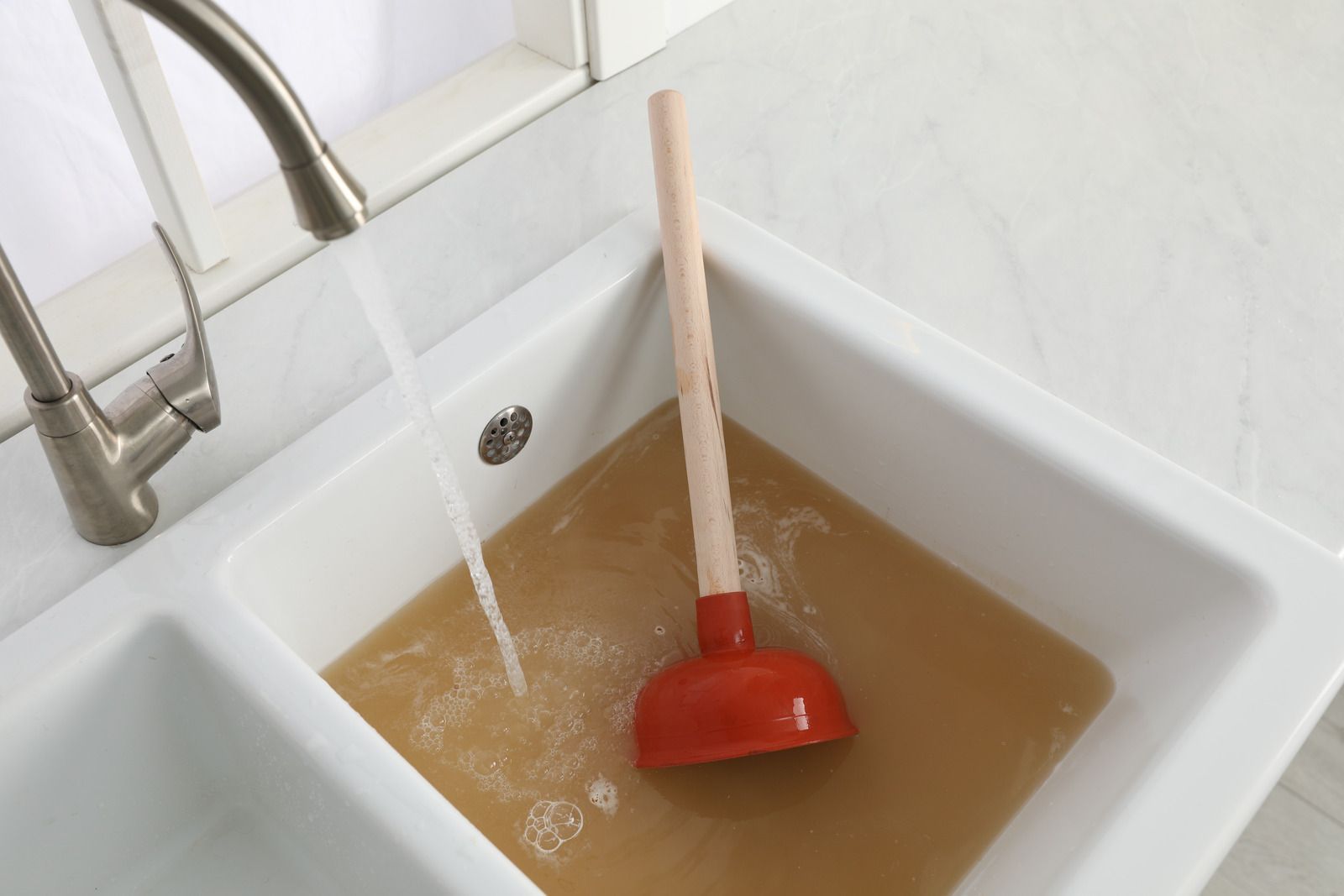 Red plunger in a sink filled with cloudy brown water under a running faucet.