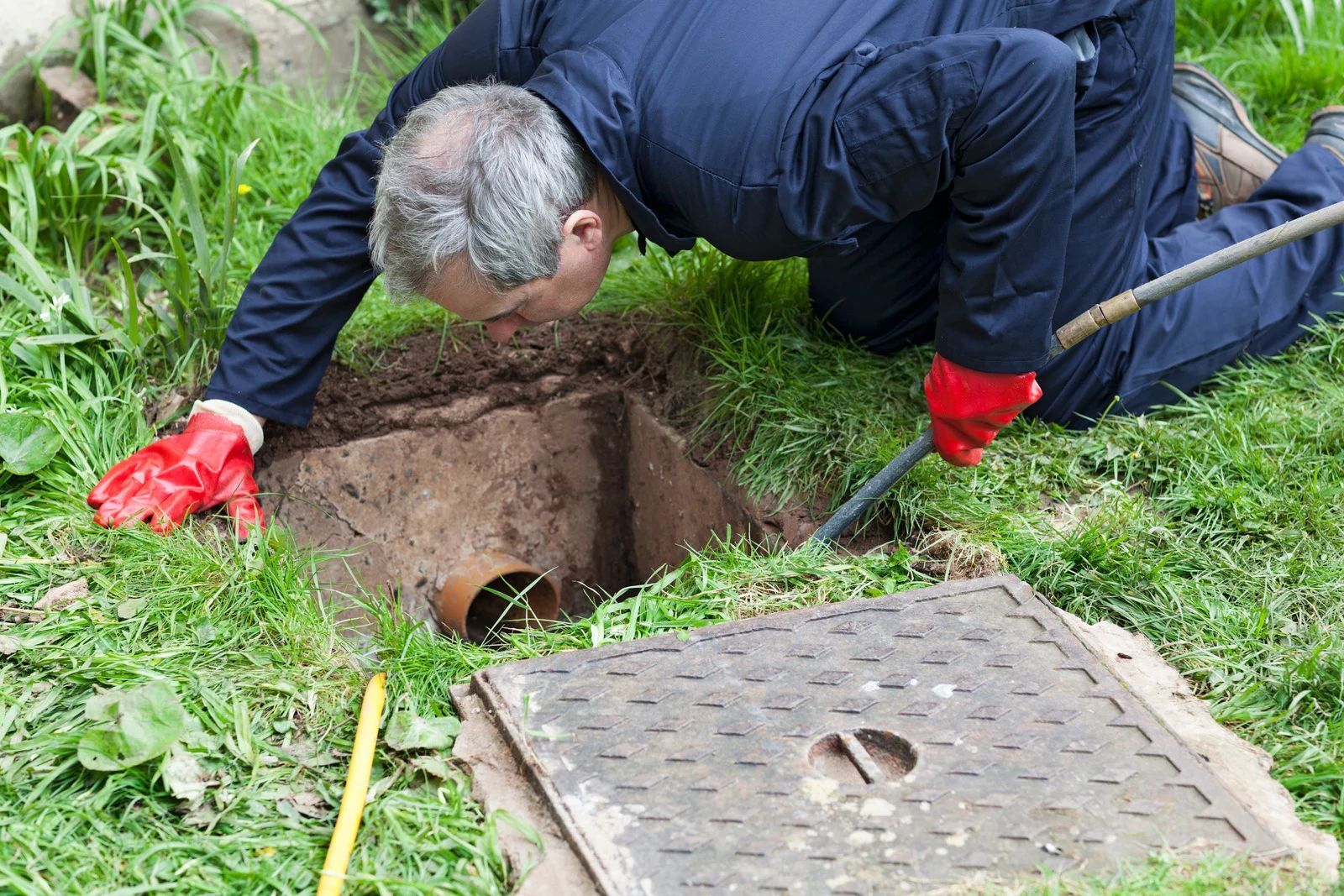 Worker in red gloves inspecting a hole in the grass near an open manhole cover.