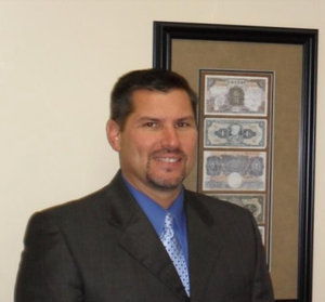 Man in dark suit and blue tie standing beside framed currency display on a wall