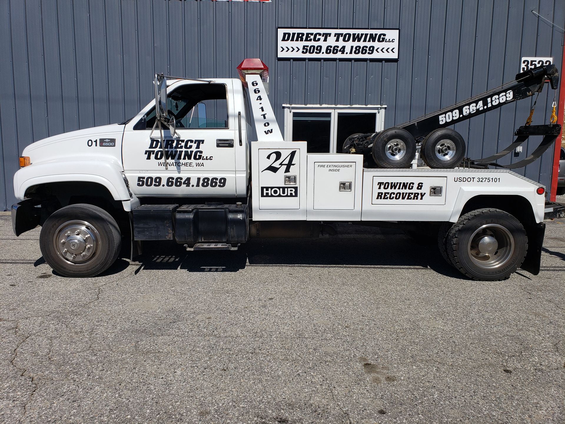 White tow truck with company decals parked on gravel beside a metal building.