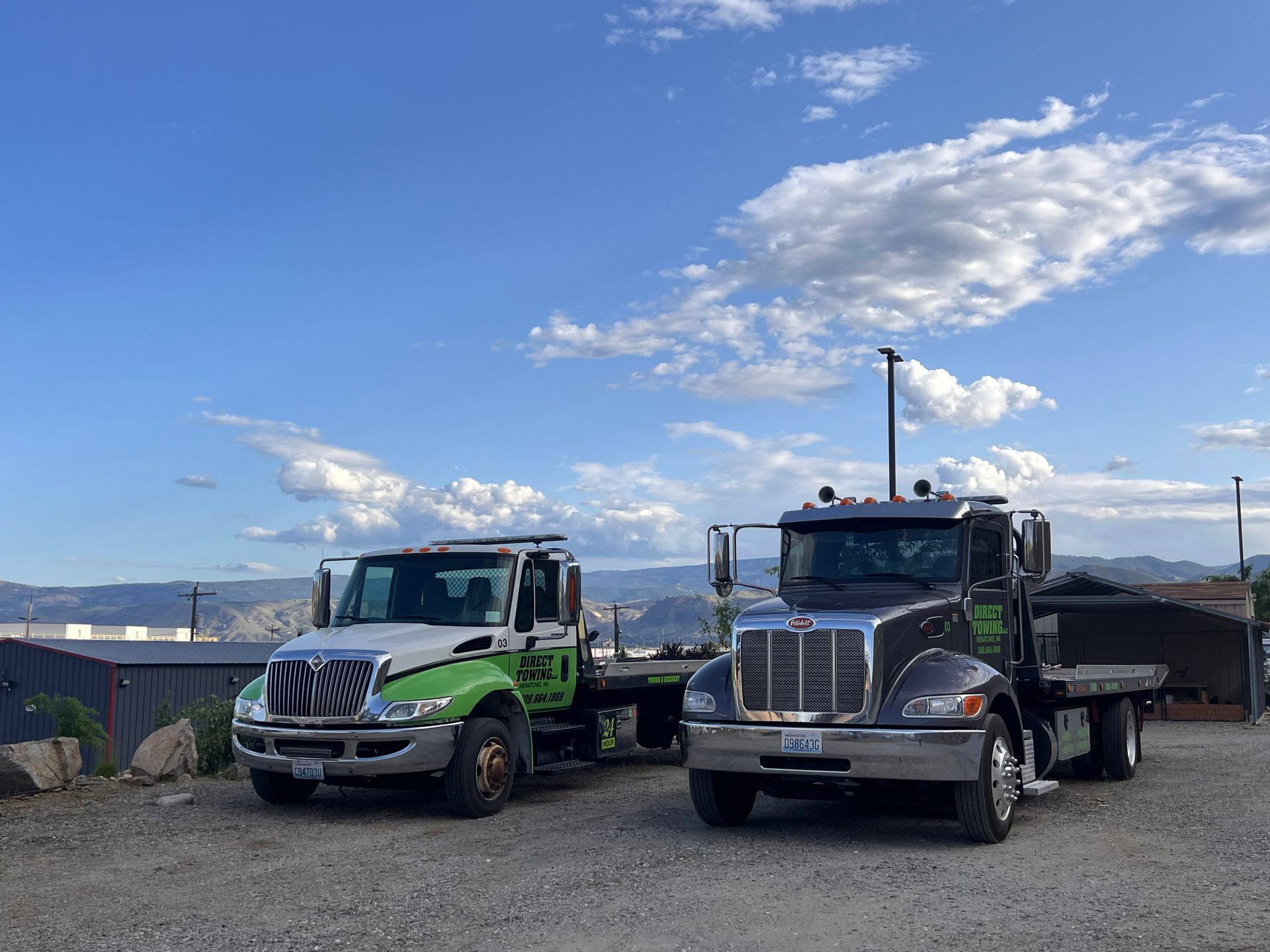 Two tow trucks parked side by side on a gravel lot under a blue sky with clouds
