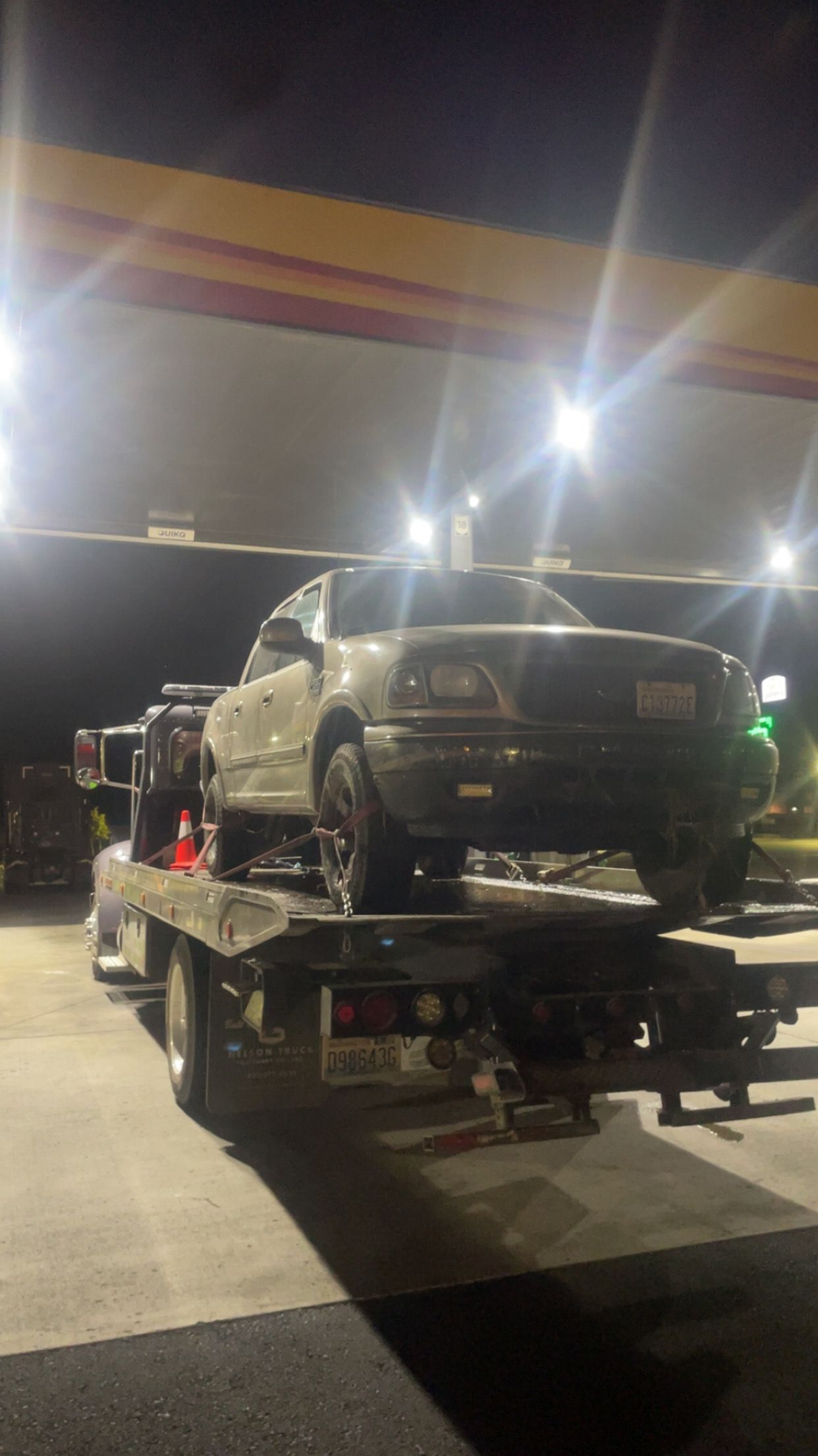 Two cars loaded on a tow truck at a lit gas station at night