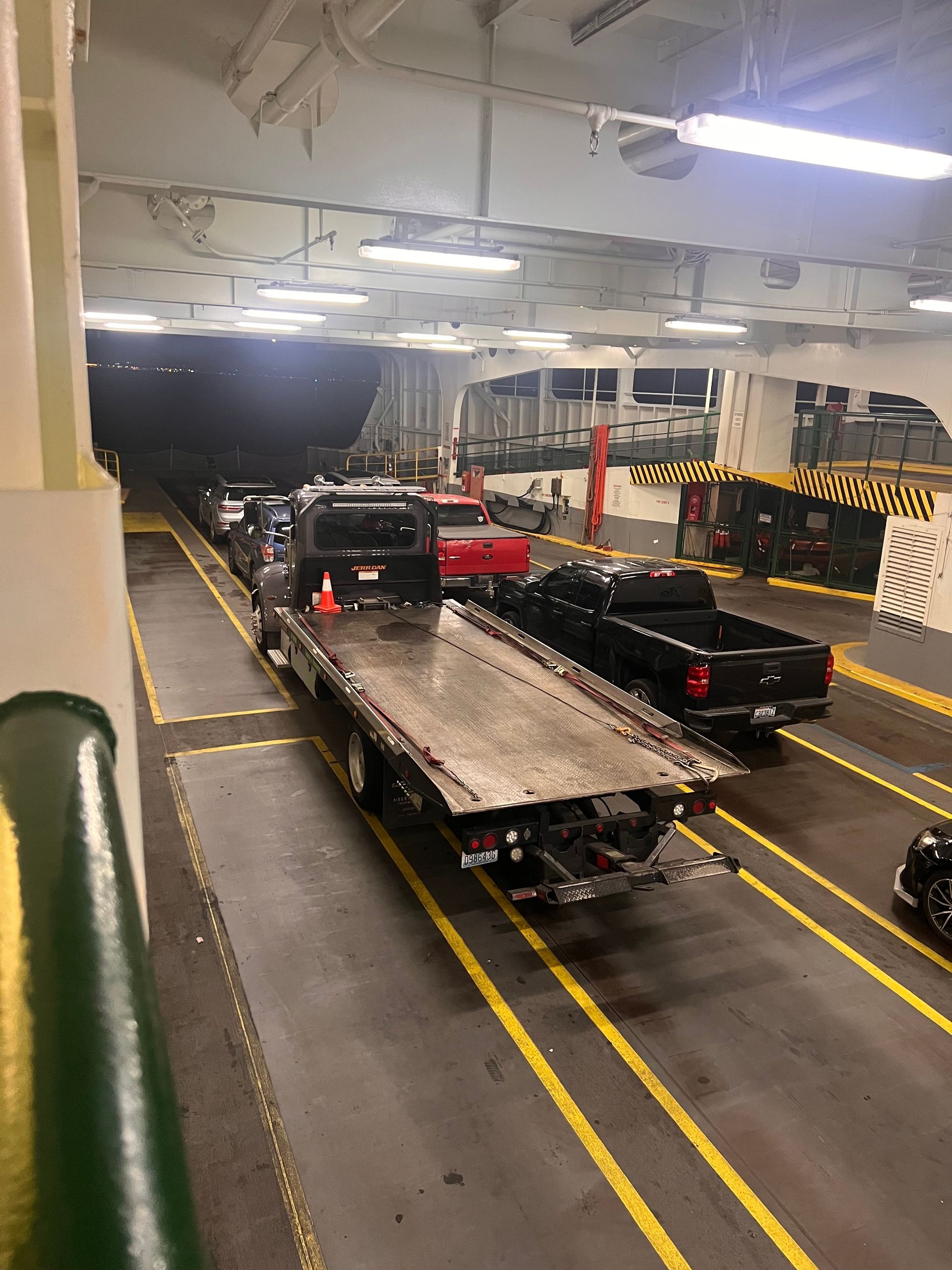 Vehicles on a ferry car deck, including a flatbed truck and black pickups, under bright overhead lights.