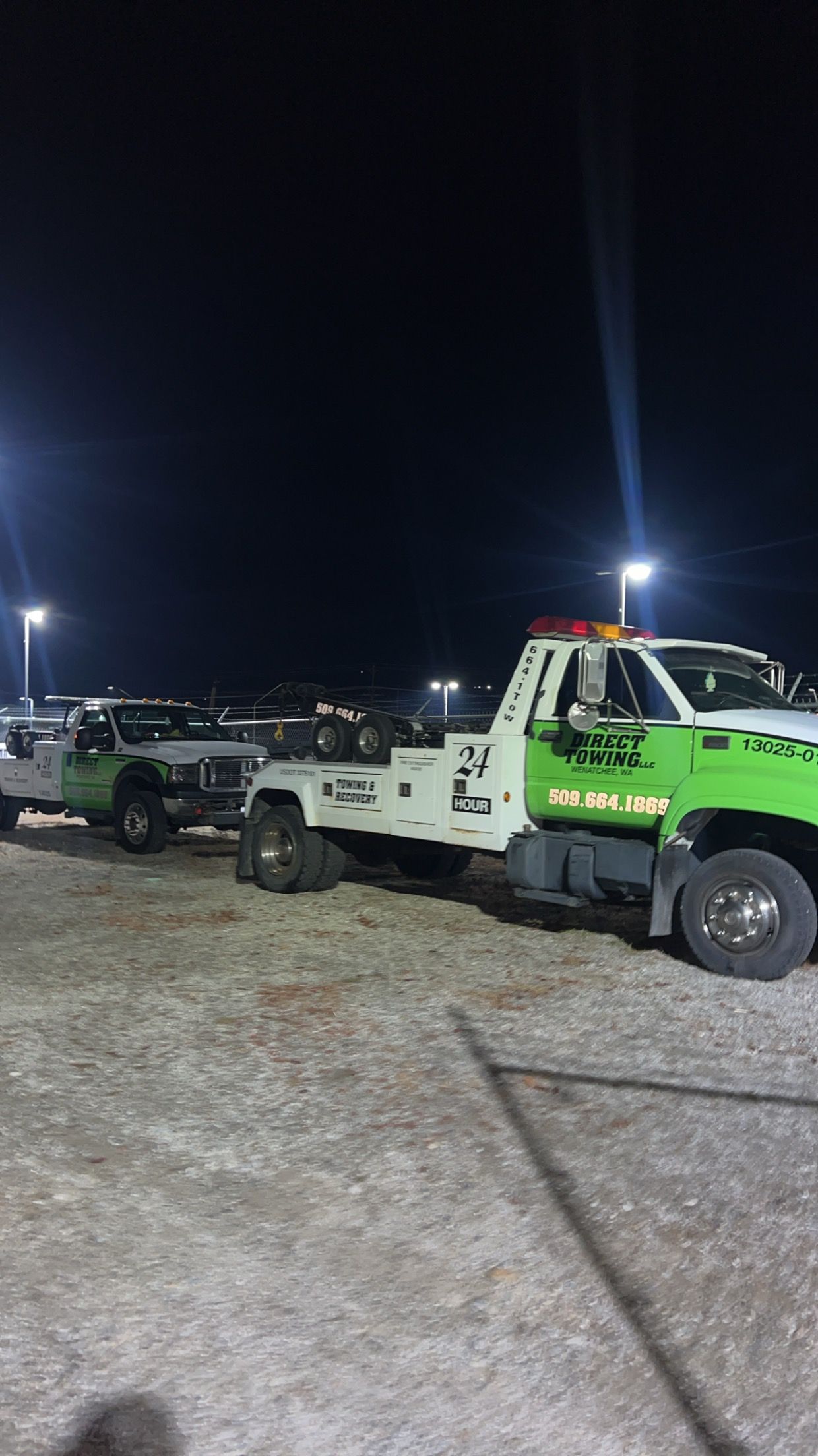 Tow trucks lined up at night in a gravel lot, including a green and white truck in the foreground.