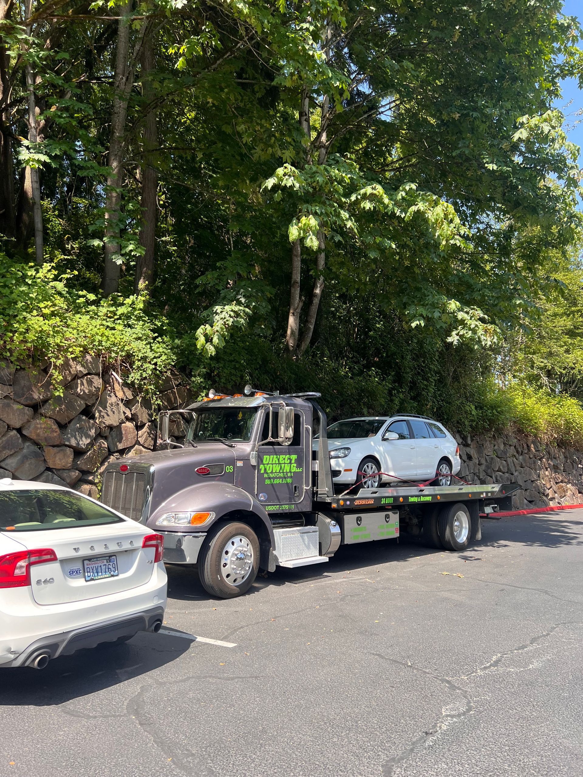 Tow truck hauling a white car on a flatbed beside a wooded roadside, with another parked car nearby