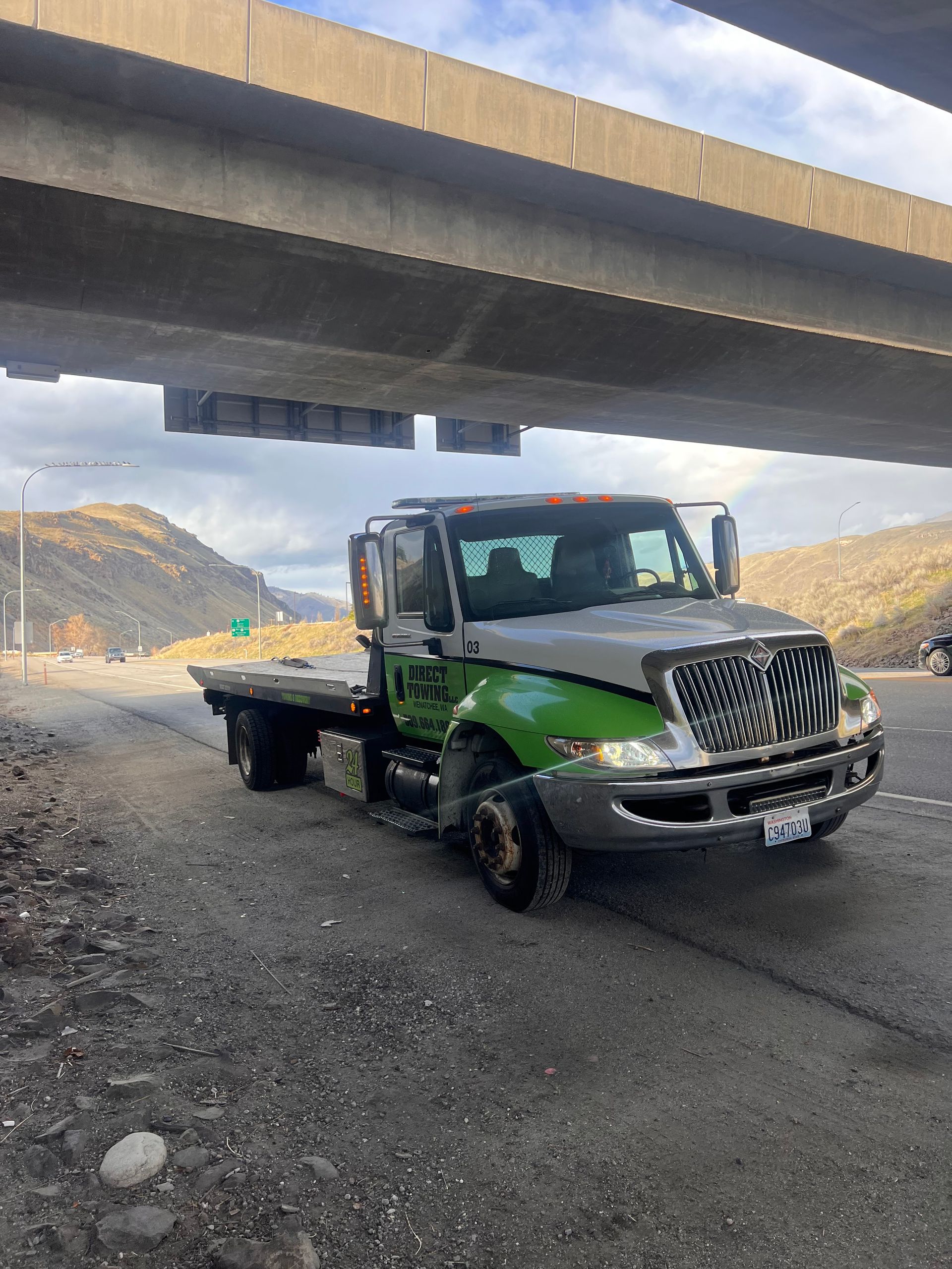 Green tow truck parked under a highway overpass on a gravel road in a mountainous area