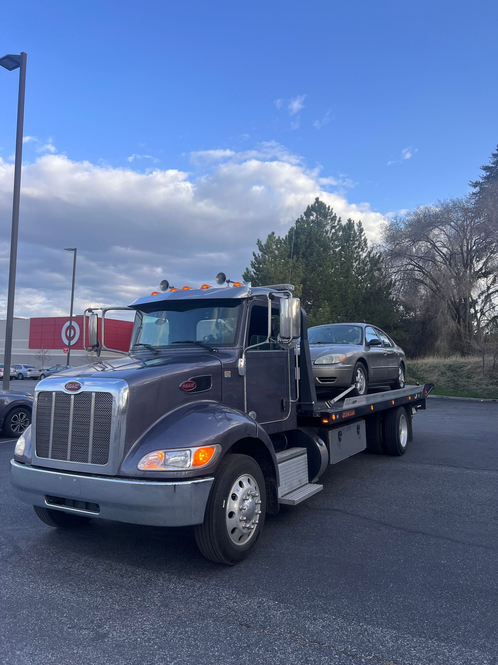 Tow truck carrying a black sedan on a flatbed in a parking lot under a blue sky