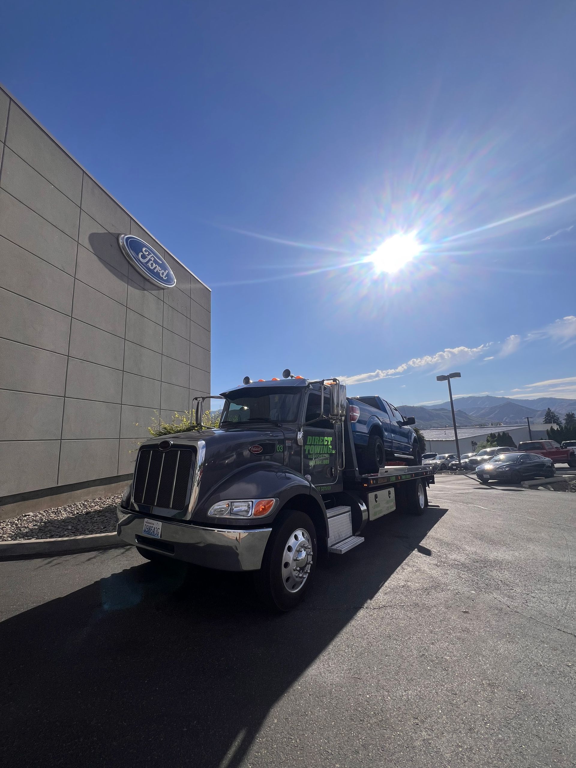 Black tow truck parked in a sunny lot beside a large building, with mountains in the distance