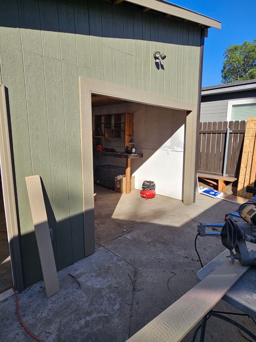 Open garage with green siding, concrete driveway, and tools inside a small backyard area.