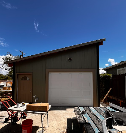 Small detached garage with a white door, green siding, and a slanted roof under a blue sky.