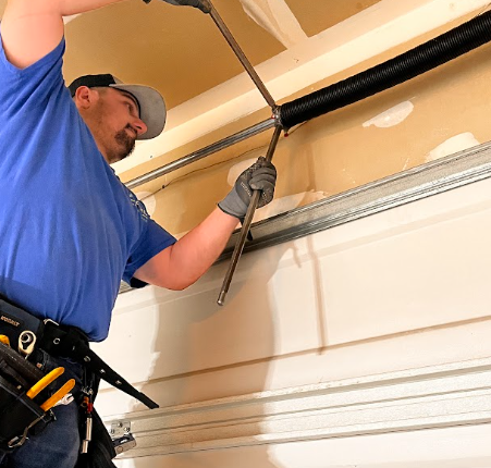 Technician in blue shirt adjusts a garage door spring with a long metal tool.