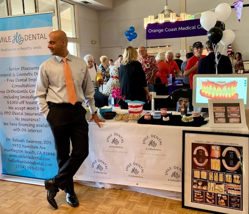 Dr. Swaroop in white shirt and orange tie at a busy indoor market booth with balloons and dessert displays