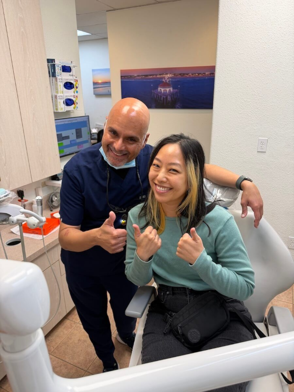 Two smiling people in a clinic, giving thumbs up while seated beside dental equipment.