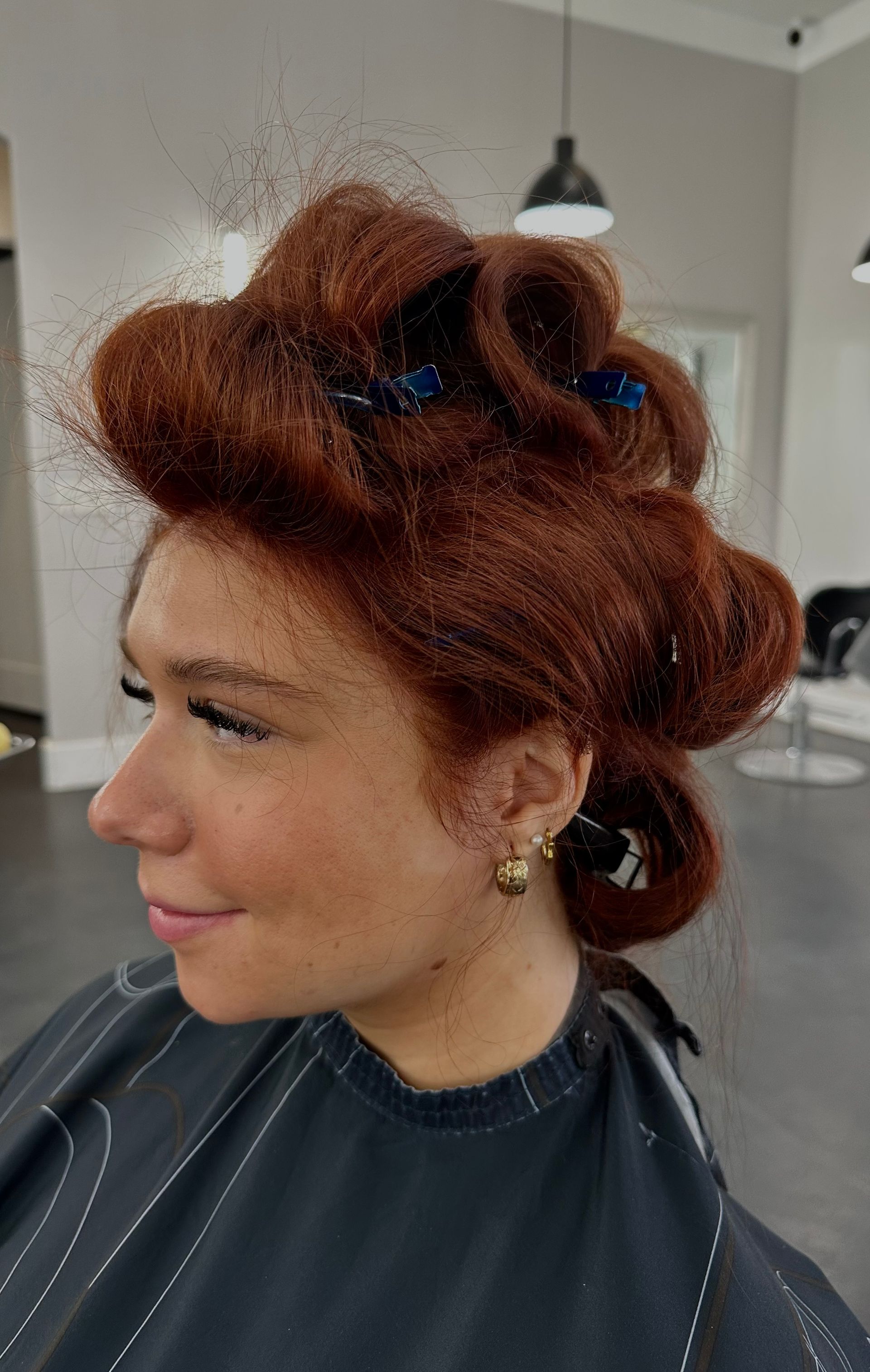 Woman with red hair in curlers at a salon, smiling.