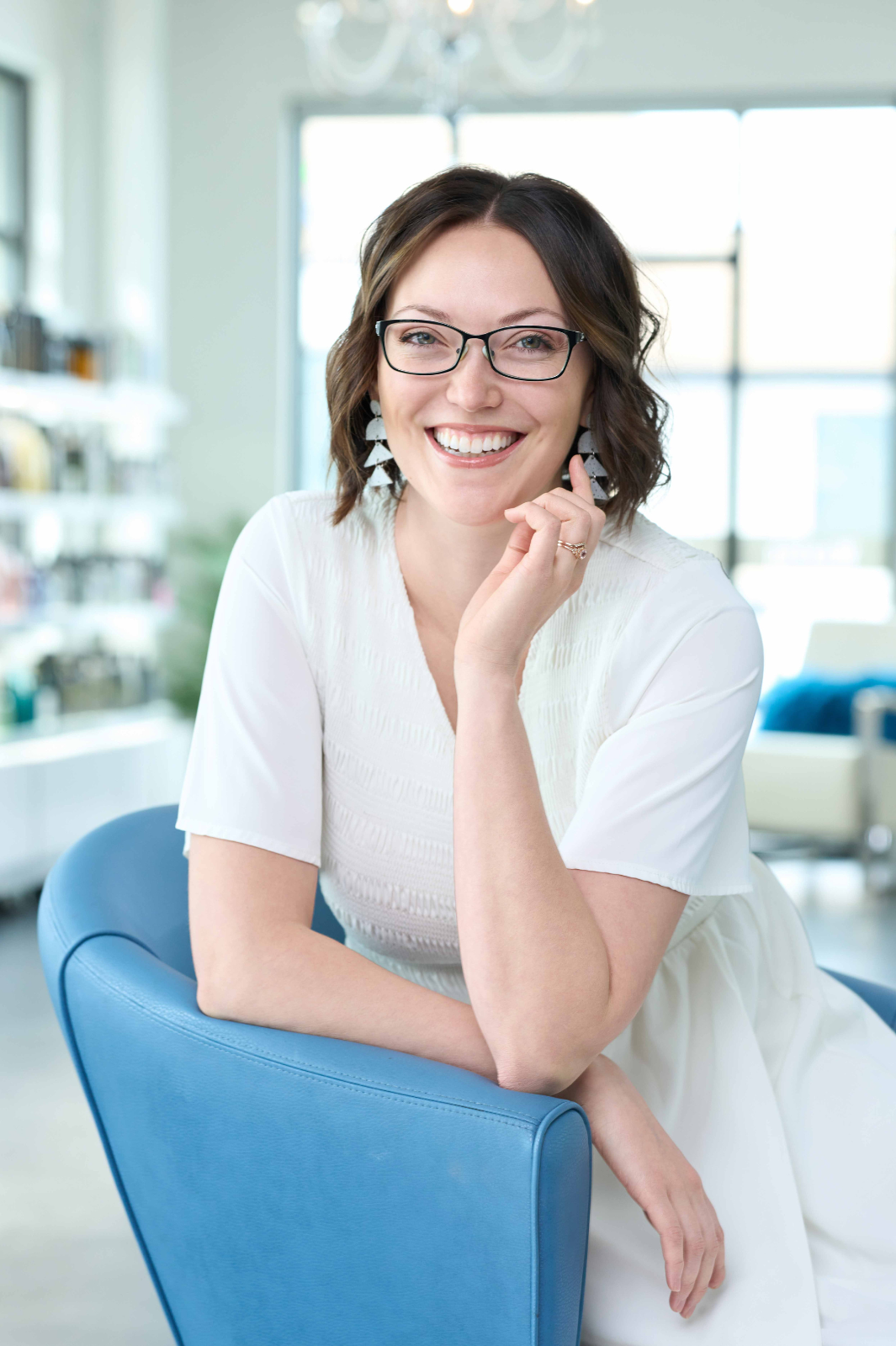 A woman wearing glasses and a white dress is sitting in a blue chair.