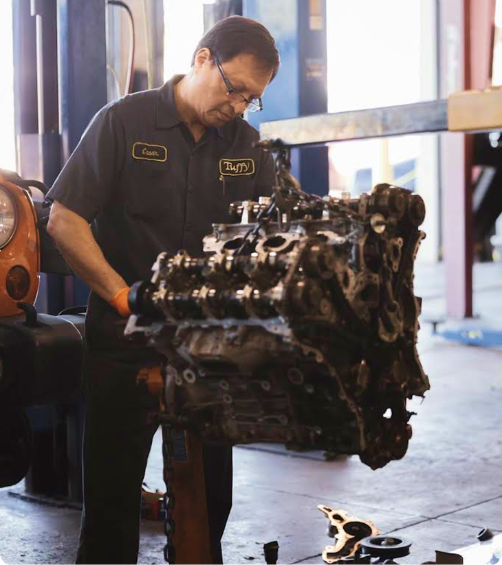 Mechanic examining a car engine in a garage. He is wearing glasses, dark clothes and is focused on the engine.