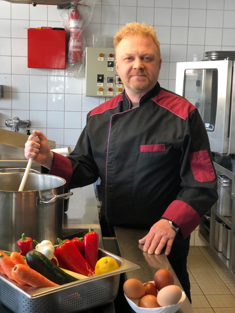 Ein Mann in Kochuniform steht mit einer Schöpfkelle in der Hand in einer Küche.