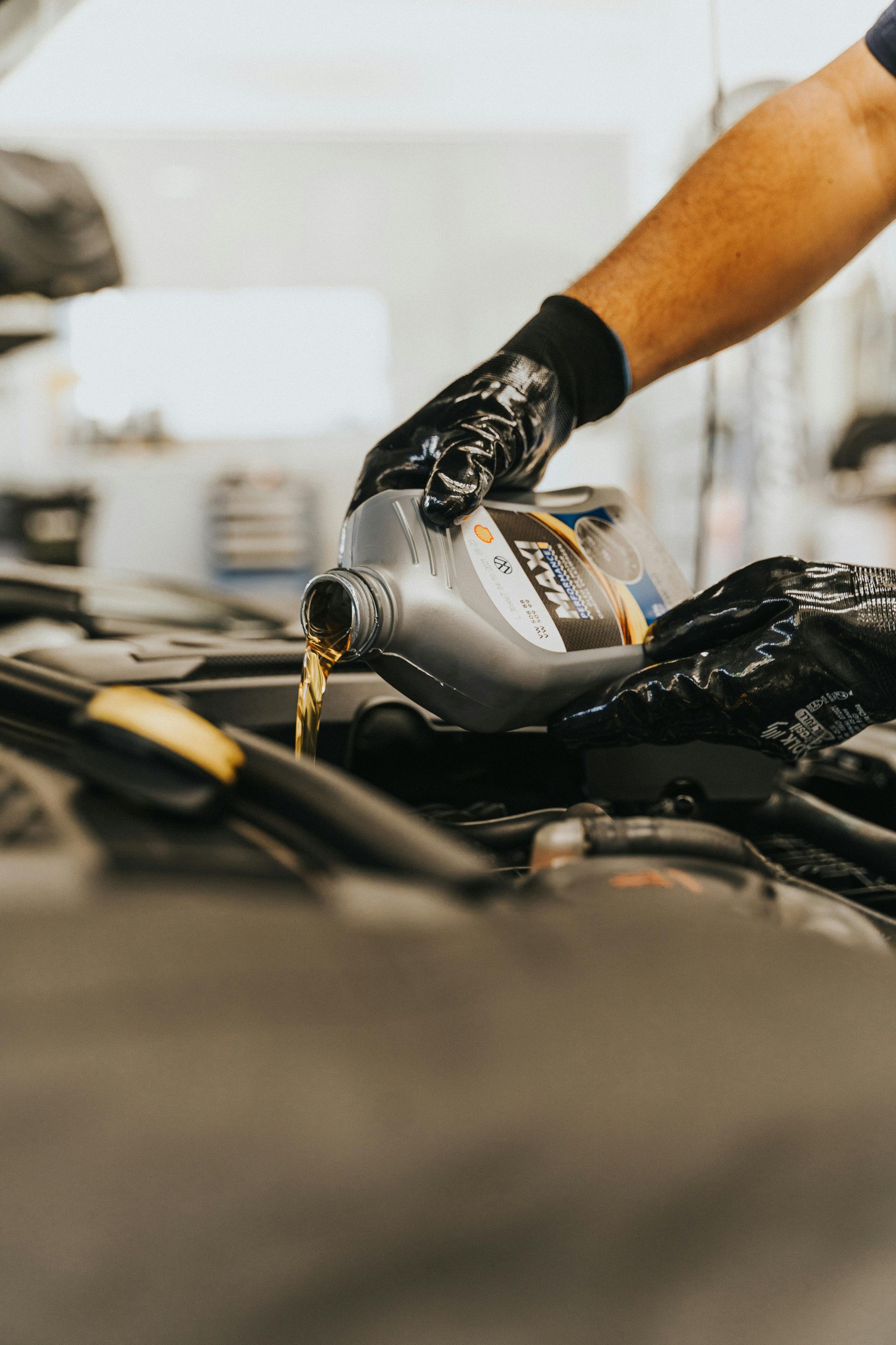 Gloved hands pouring motor oil into an engine bay from a gray bottle