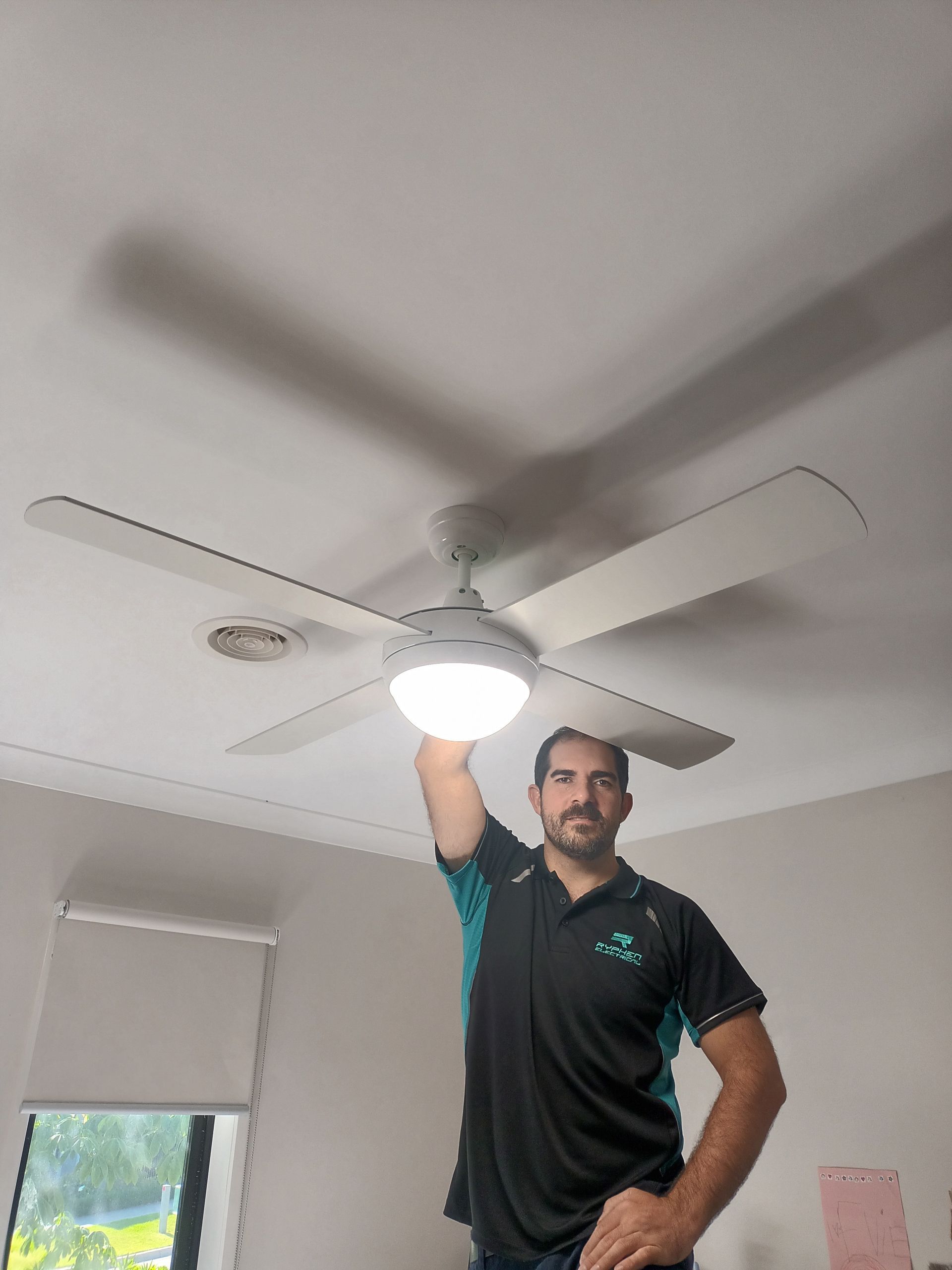 Man standing near a ceiling fan, reaching up to the light fixture. Inside a room with white ceiling and walls. — Ryphen Electrical in Highton, VIC
