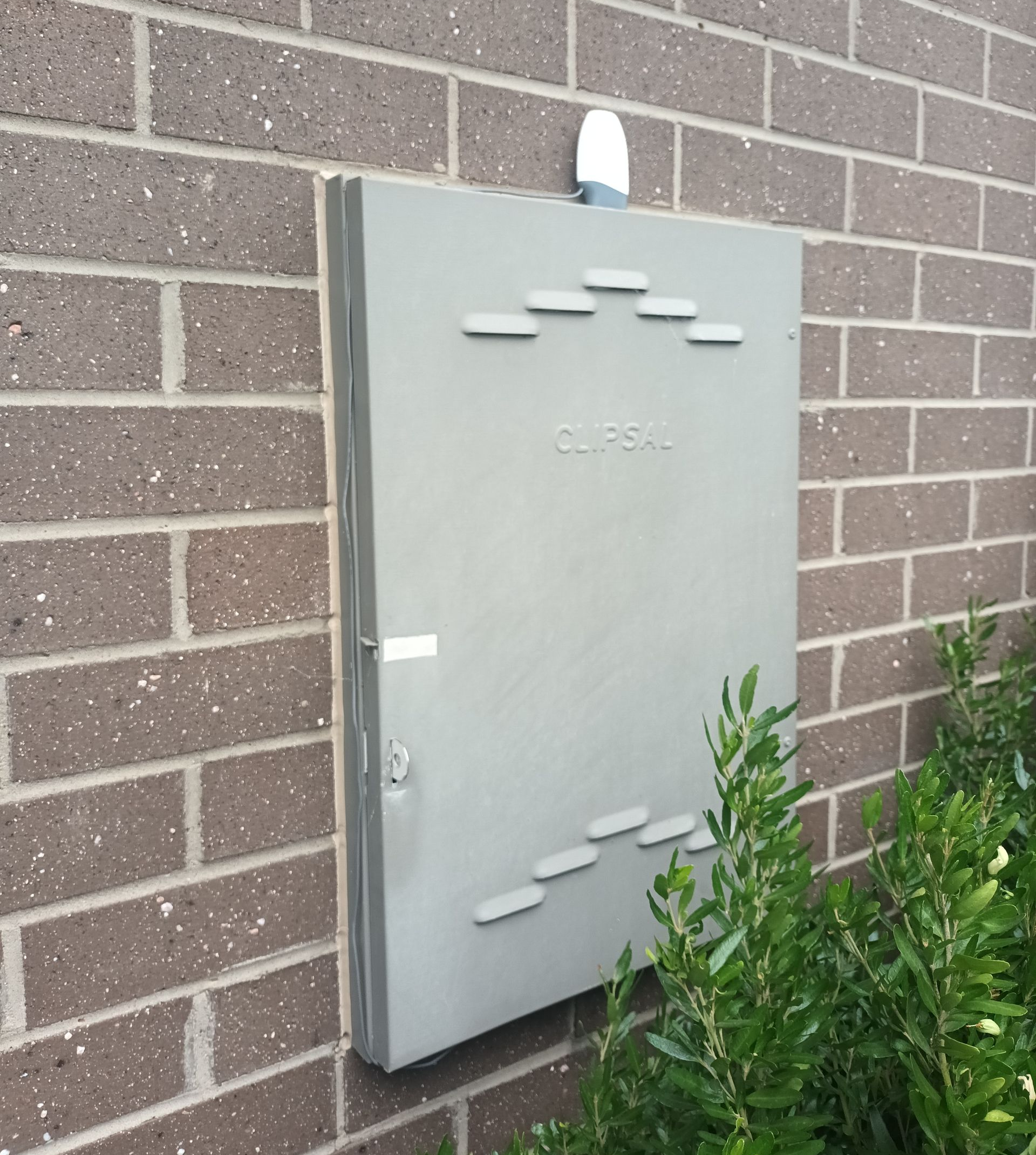 Gray rectangular utility box on a brick wall with a white fixture above it, foliage in the foreground. — Ryphen Electrical in Highton, VIC