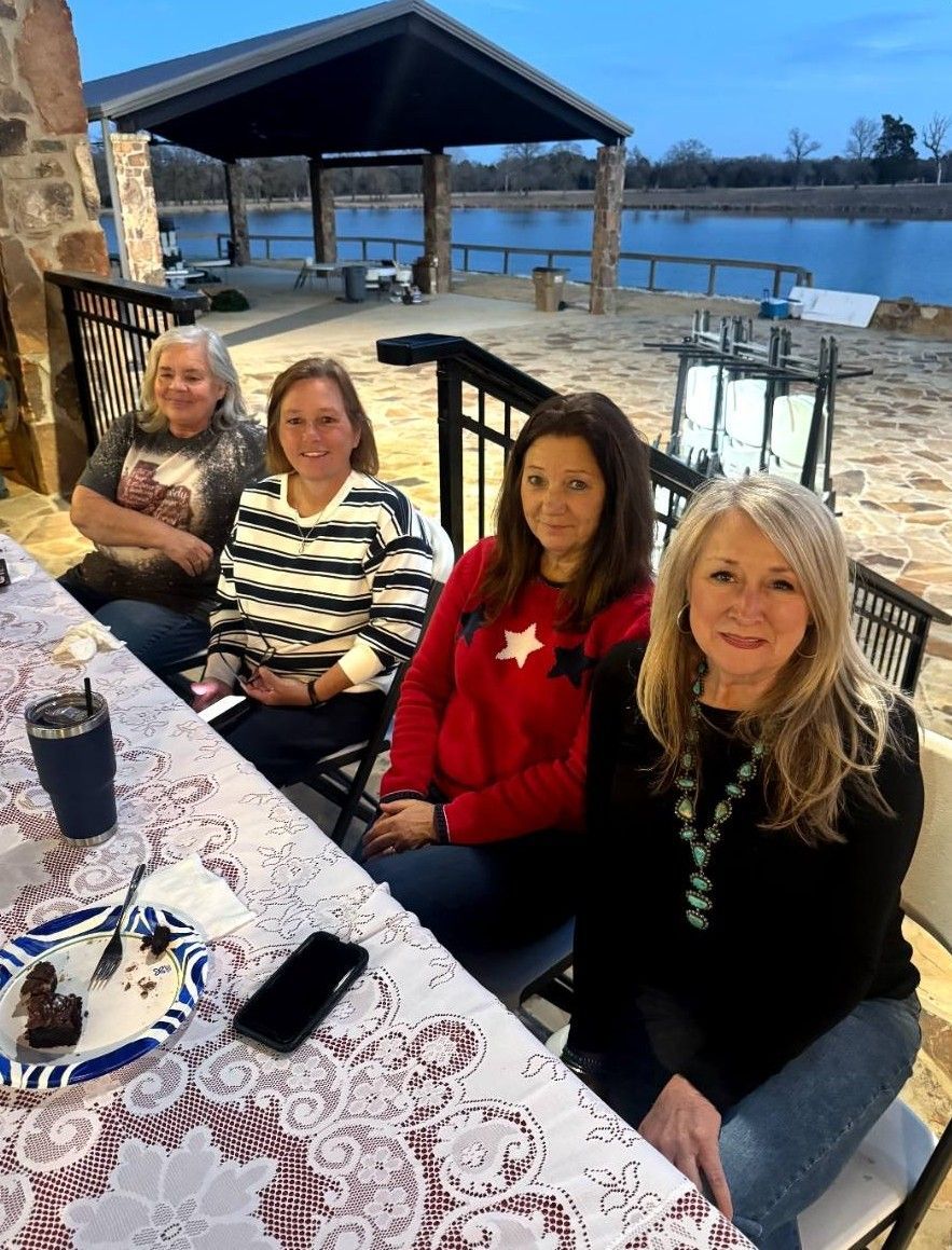 Four people seated at an outdoor lakeside table, smiling beside a stone patio and pavilion.