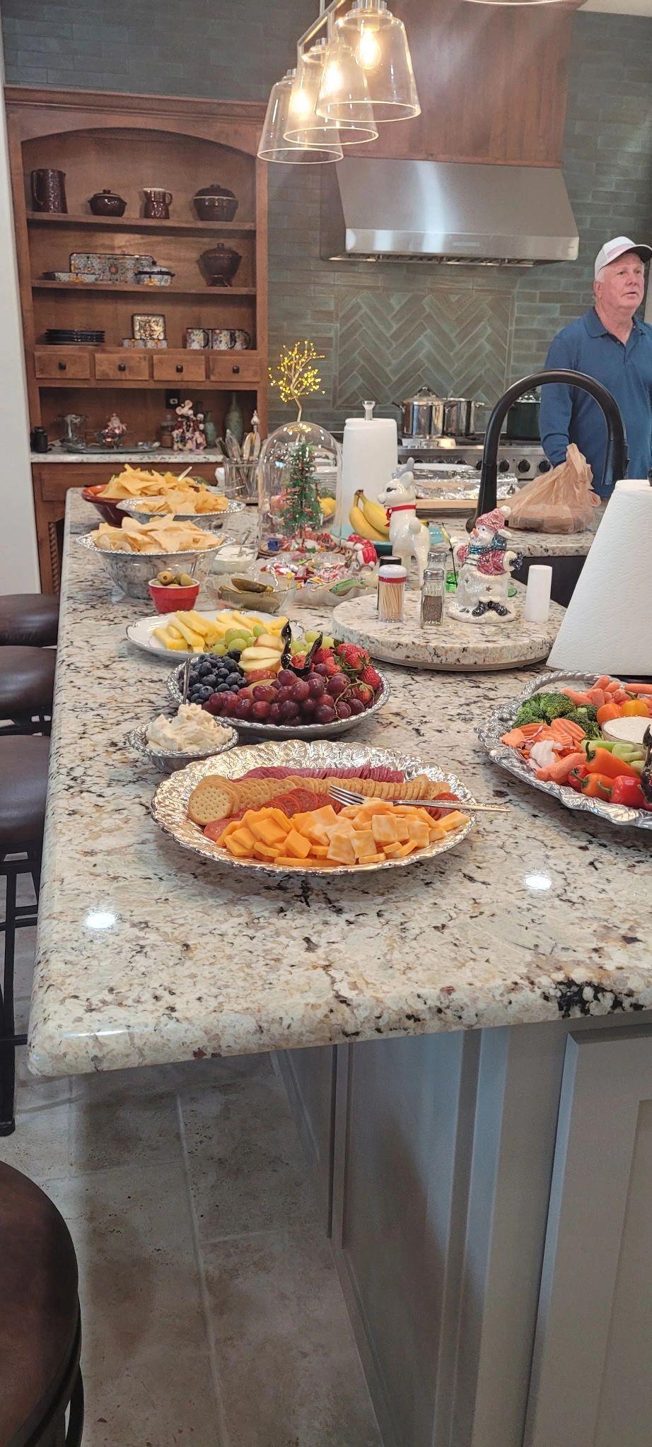 Kitchen counter covered with trays of assorted desserts and fruit, with a person standing in the background.