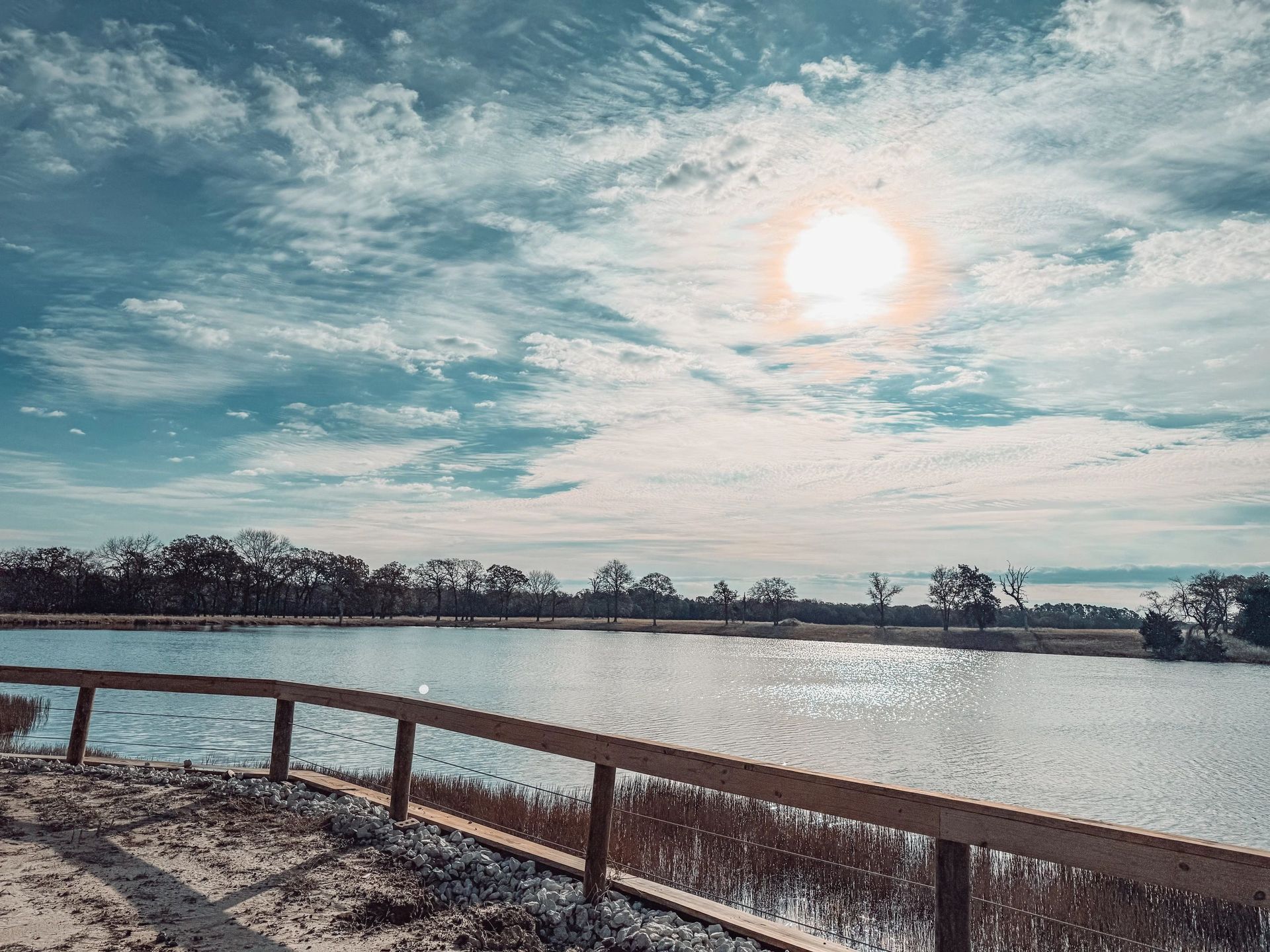 Sunlit winter lake with bare trees and a wooden fence along the shore