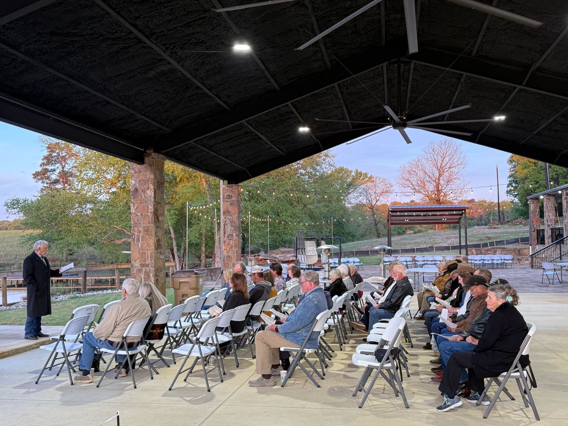 Audience seated under pavilion at outdoor event, with autumn trees and stage in background.