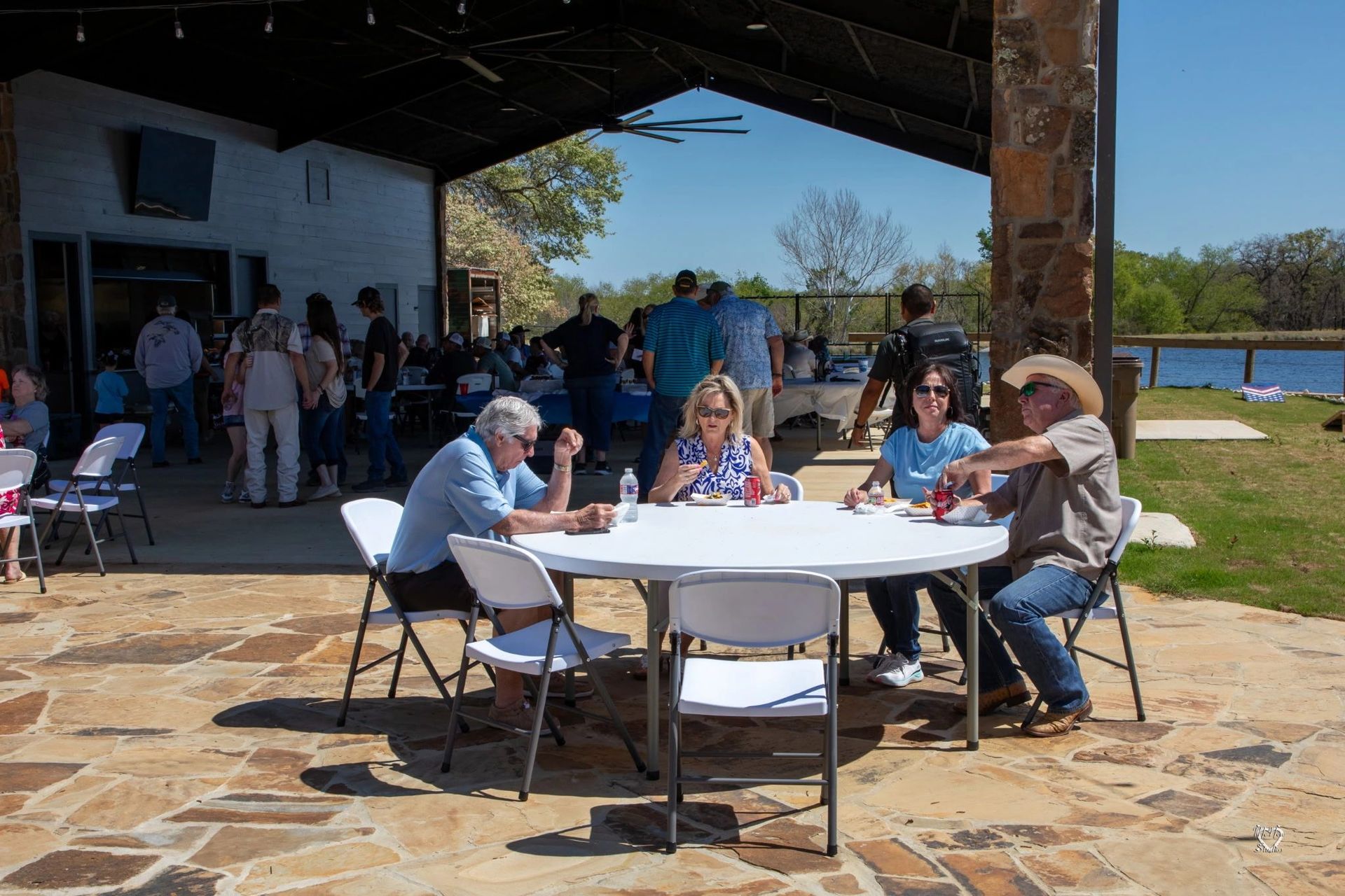 People seated at tables on a stone patio under a shaded pavilion by a lake.