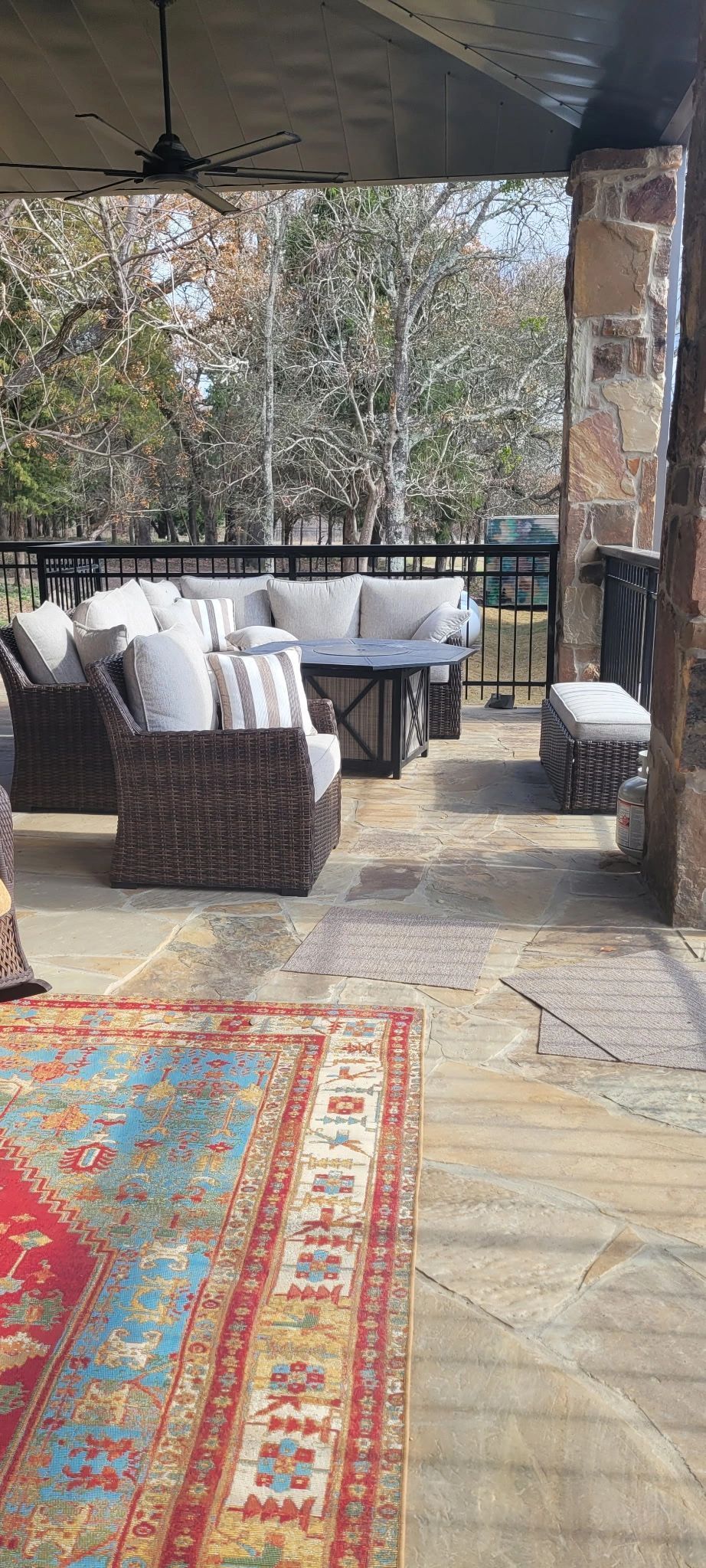 Covered patio with wicker chairs, tables, and colorful rug overlooking trees and stone floor