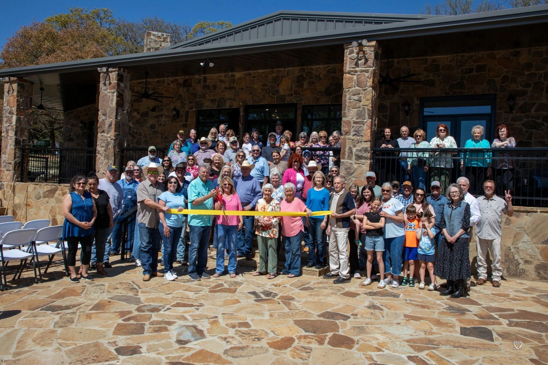 Large group posing outside a stone building, holding a ribbon at what appears to be a ribbon-cutting ceremony.