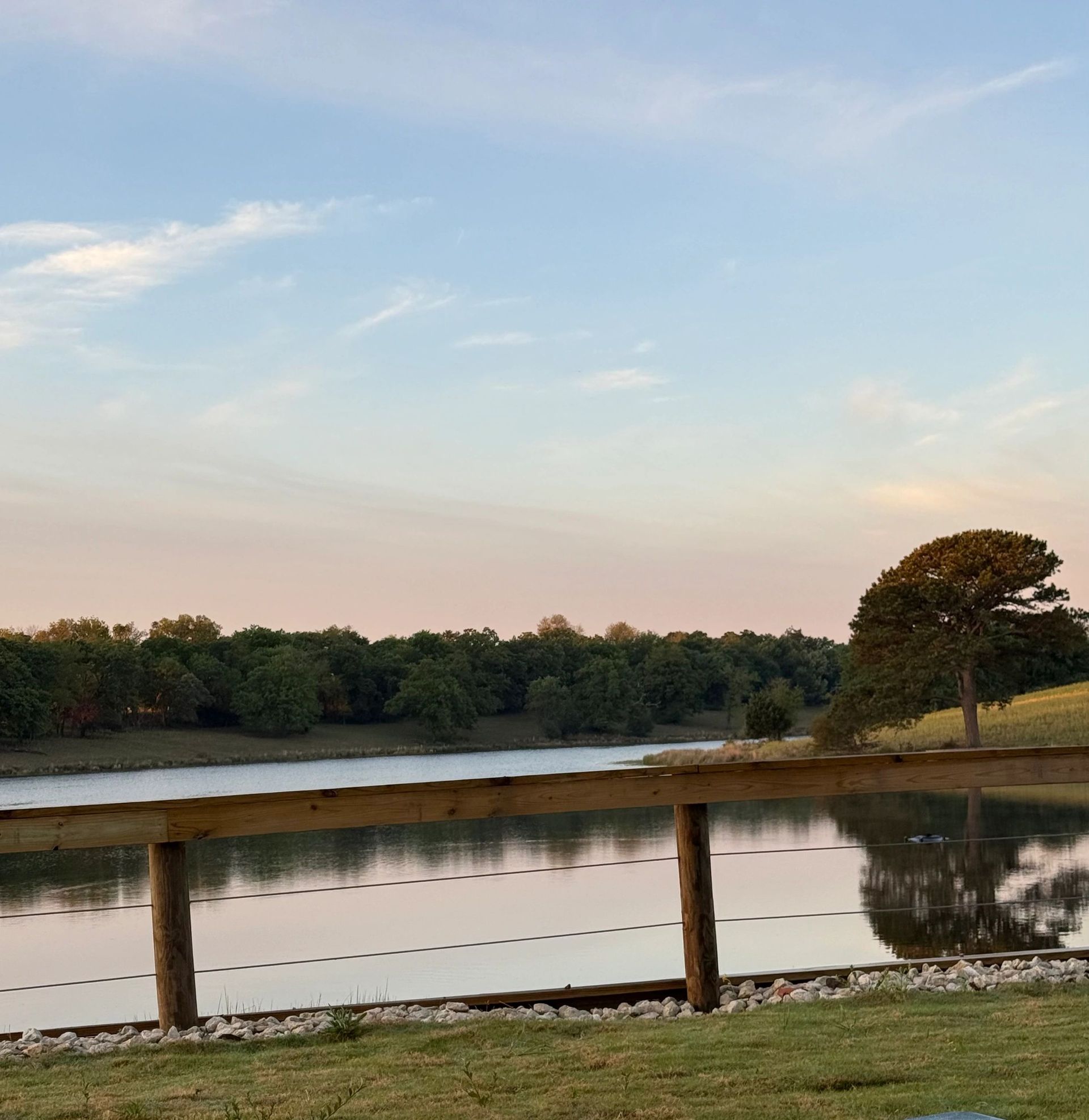 Wooden fence by a calm pond at sunset, with trees and a blue sky reflected in the water.