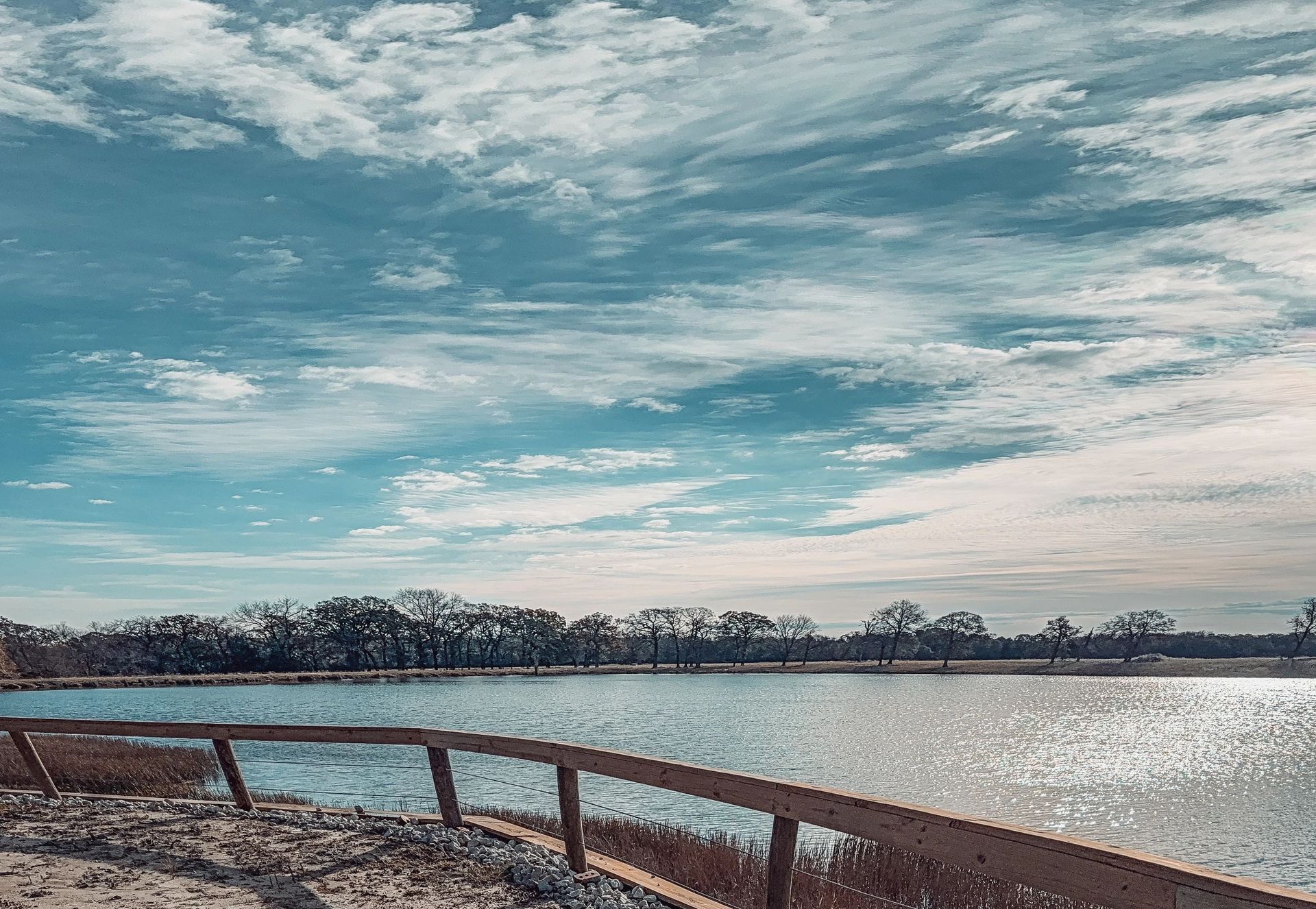 Lake and cloudy sky above a wooden fence, with bright sunlight reflecting on the water.