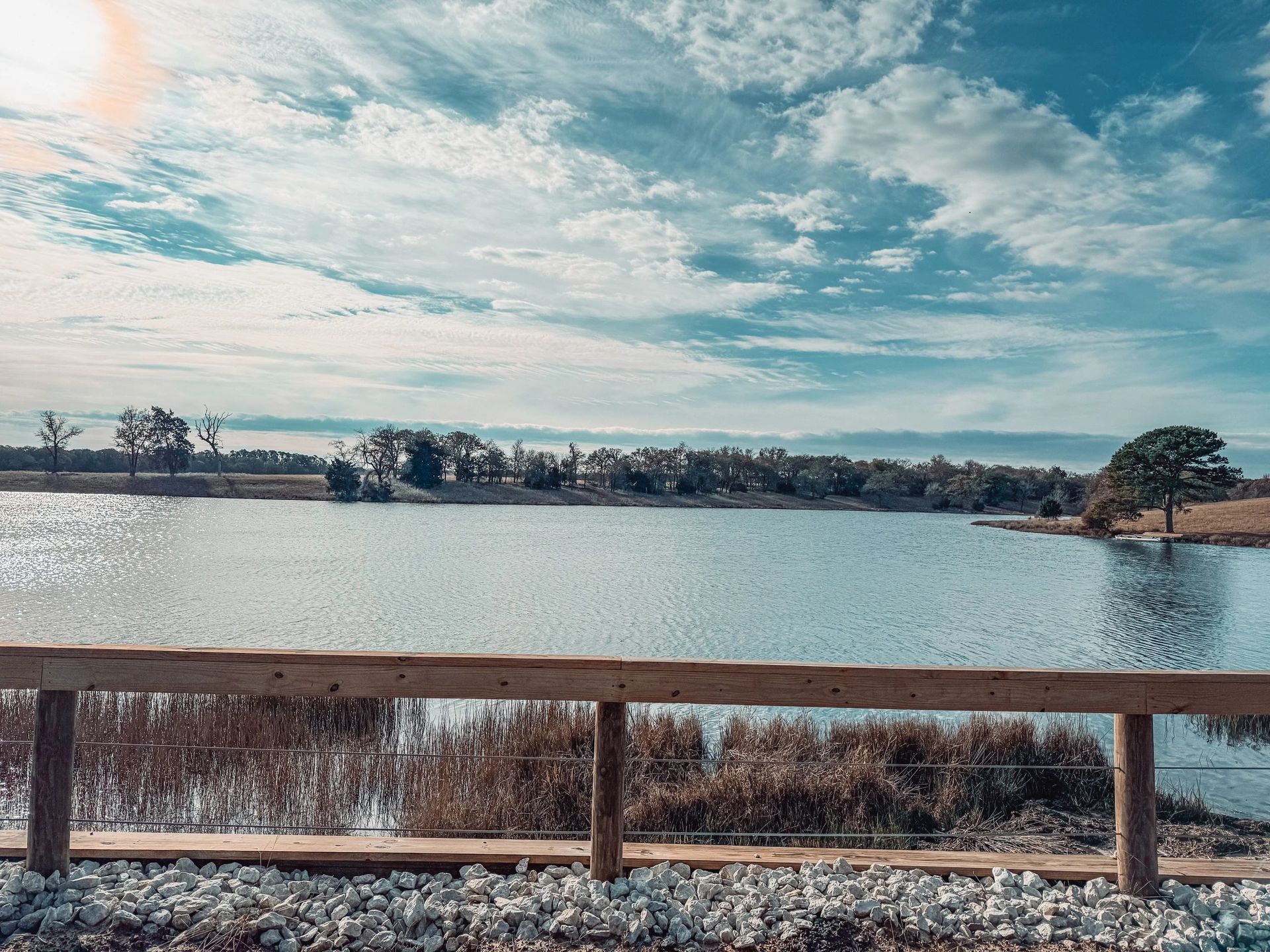 Calm lake with a wooden railing in front, under a cloudy blue sky with distant trees on the shore