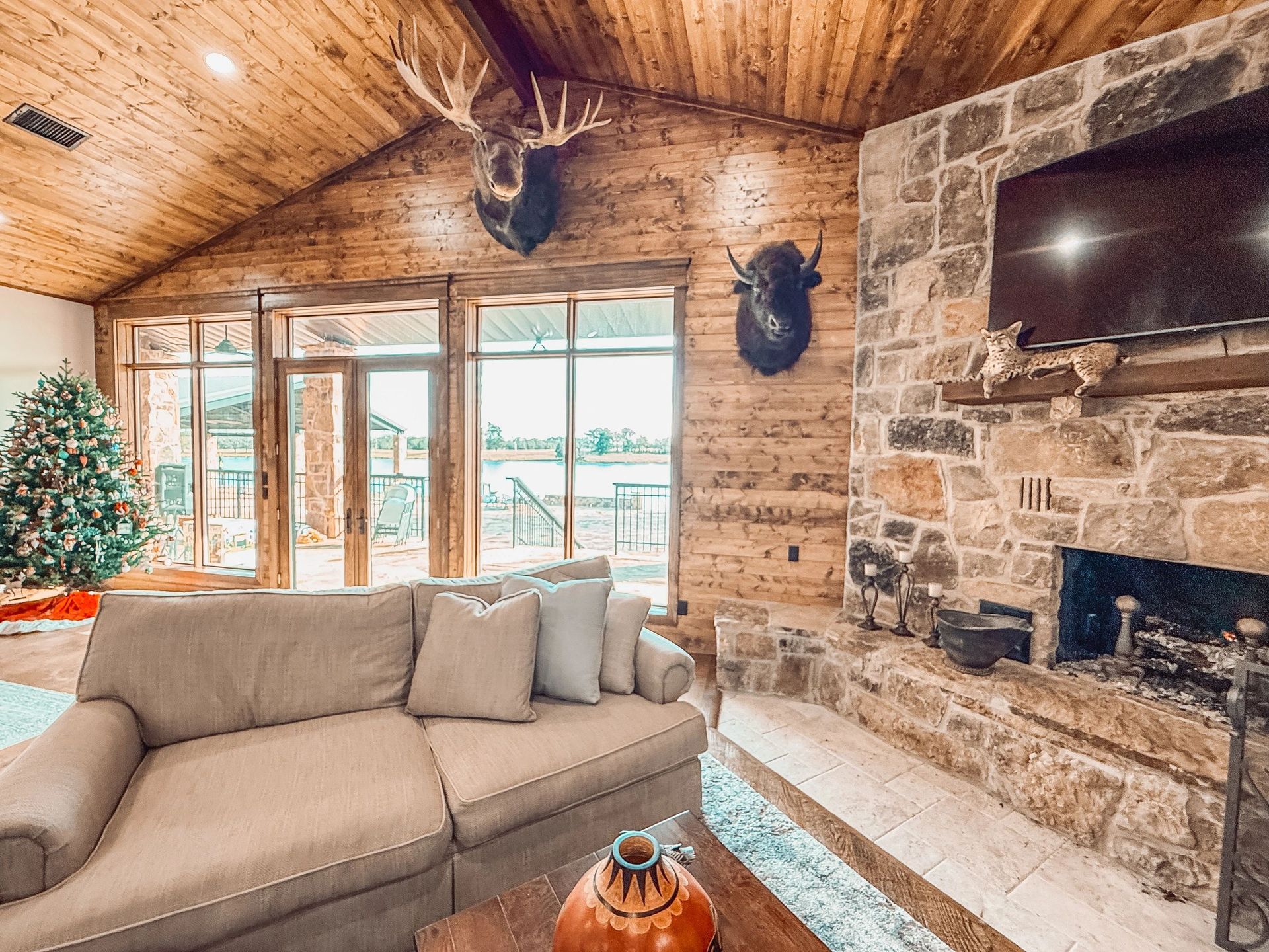 Cozy log cabin living room with stone fireplace, beige sectional sofa, TV, and Christmas tree near large windows