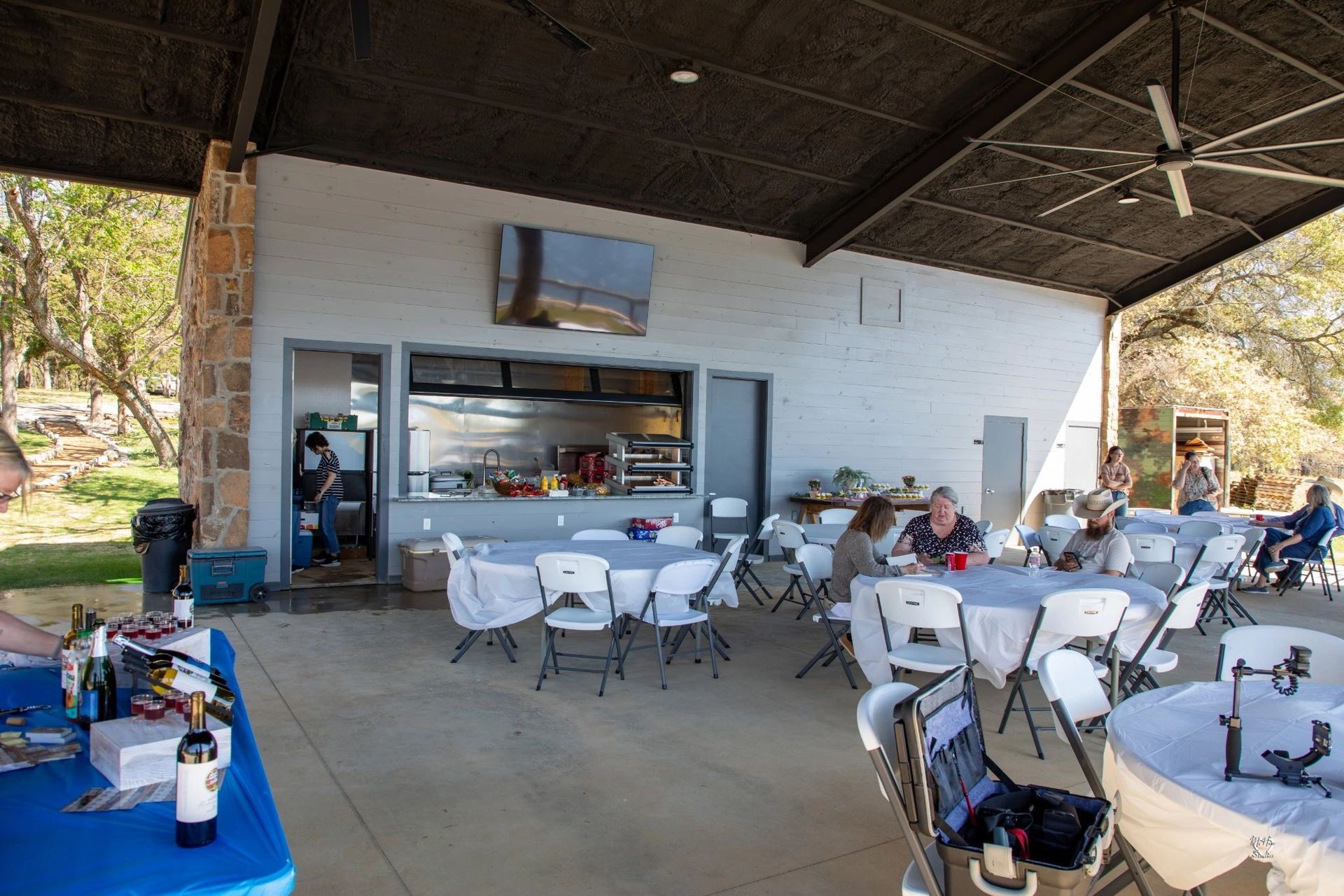 Outdoor covered picnic area with white tables and chairs, people seated, and a serving counter on the back wall