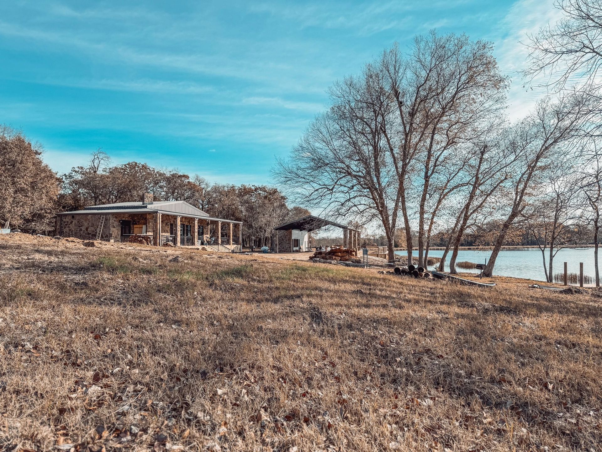 Leafy lakeside park with bare trees, a pavilion, and a calm shoreline under a blue sky