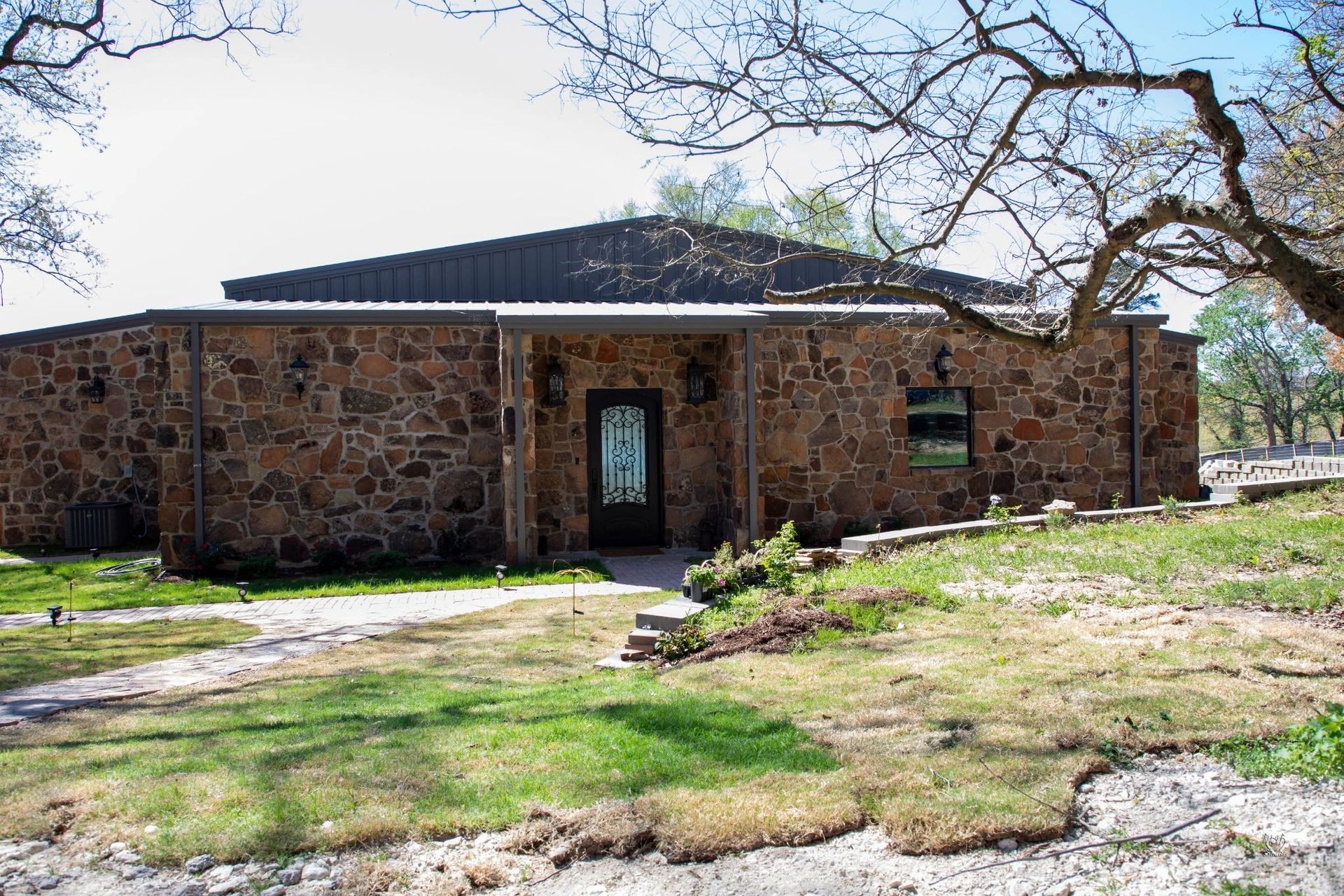 Stone house with a dark roof, central door, and bare tree branches in a grassy yard.