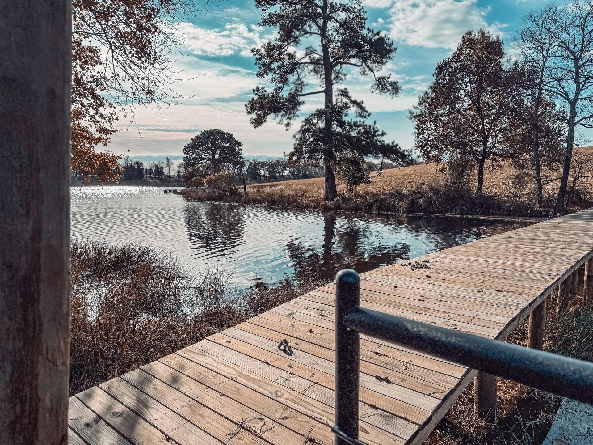 Wooden boardwalk beside a calm lake, with bare trees and a blue sky reflected in the water.