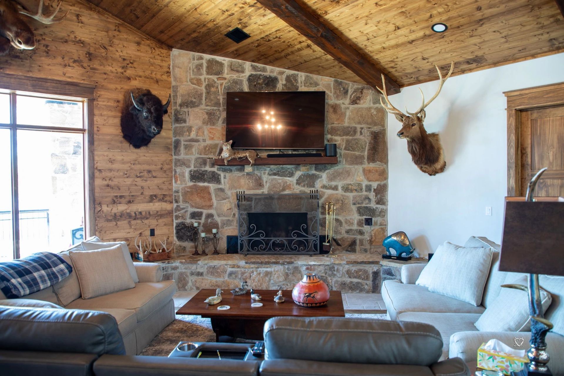 Cozy rustic living room with stone fireplace, mounted TV, antlers, and gray sofas around a coffee table