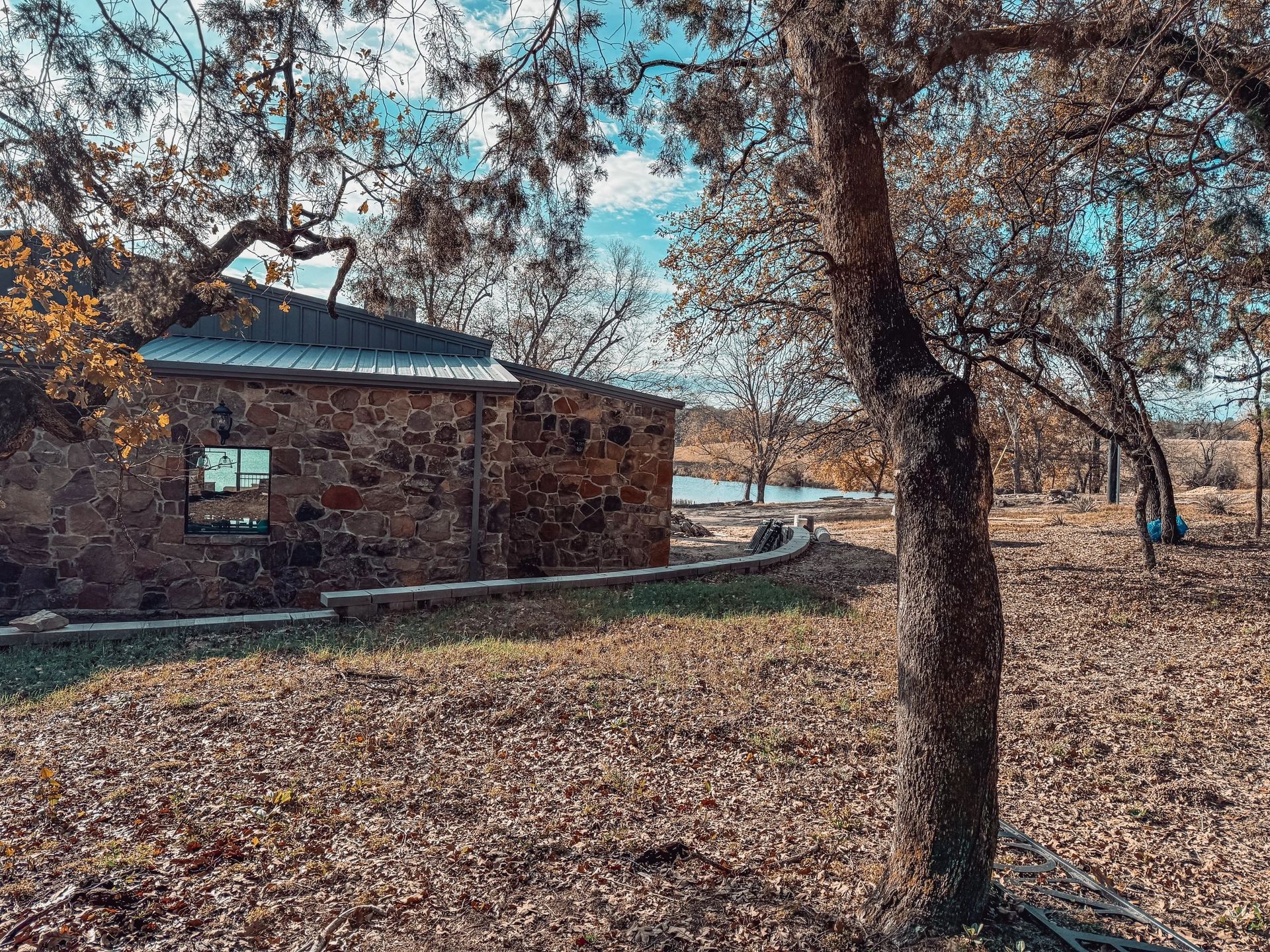 Stone cabin under leafless trees in a dry, wooded yard with a blue sky.